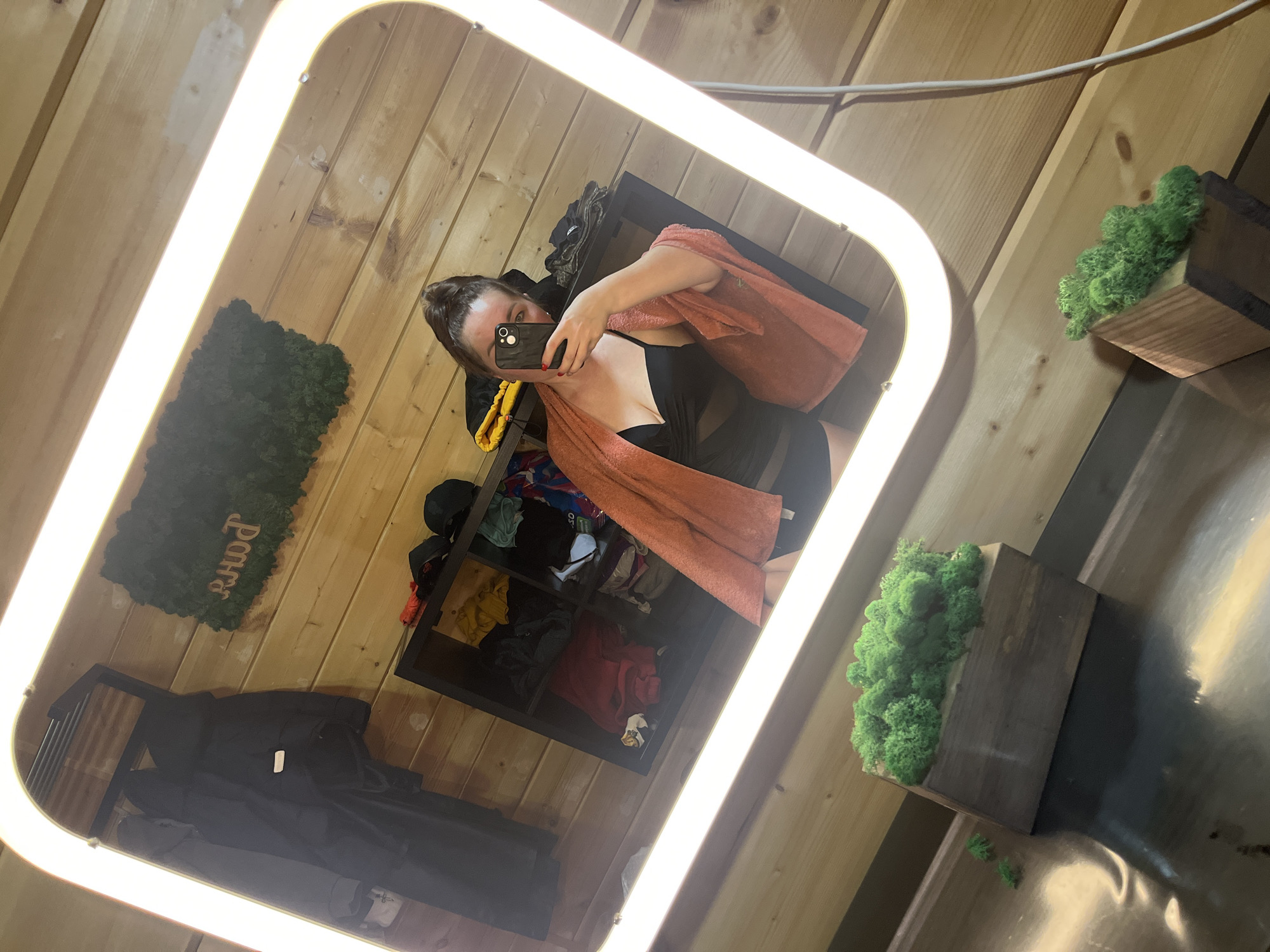 A mirror selfie is taken in a room with wooden walls and ceiling. A woman stands in front of the mirror holding a black smartphone, partially obscuring her face. She wears a black outfit and has an orange towel draped around her shoulders. Behind her is a dark shelving unit containing clothes and other items. Adjacent to the shelving unit is a dark coat hanging on a rack. On the walls are decorative elements, including a green moss panel and two small wooden shelves each holding green moss. The scene appears to be indoors, possibly a changing room. The location is Unknown, Unknown.