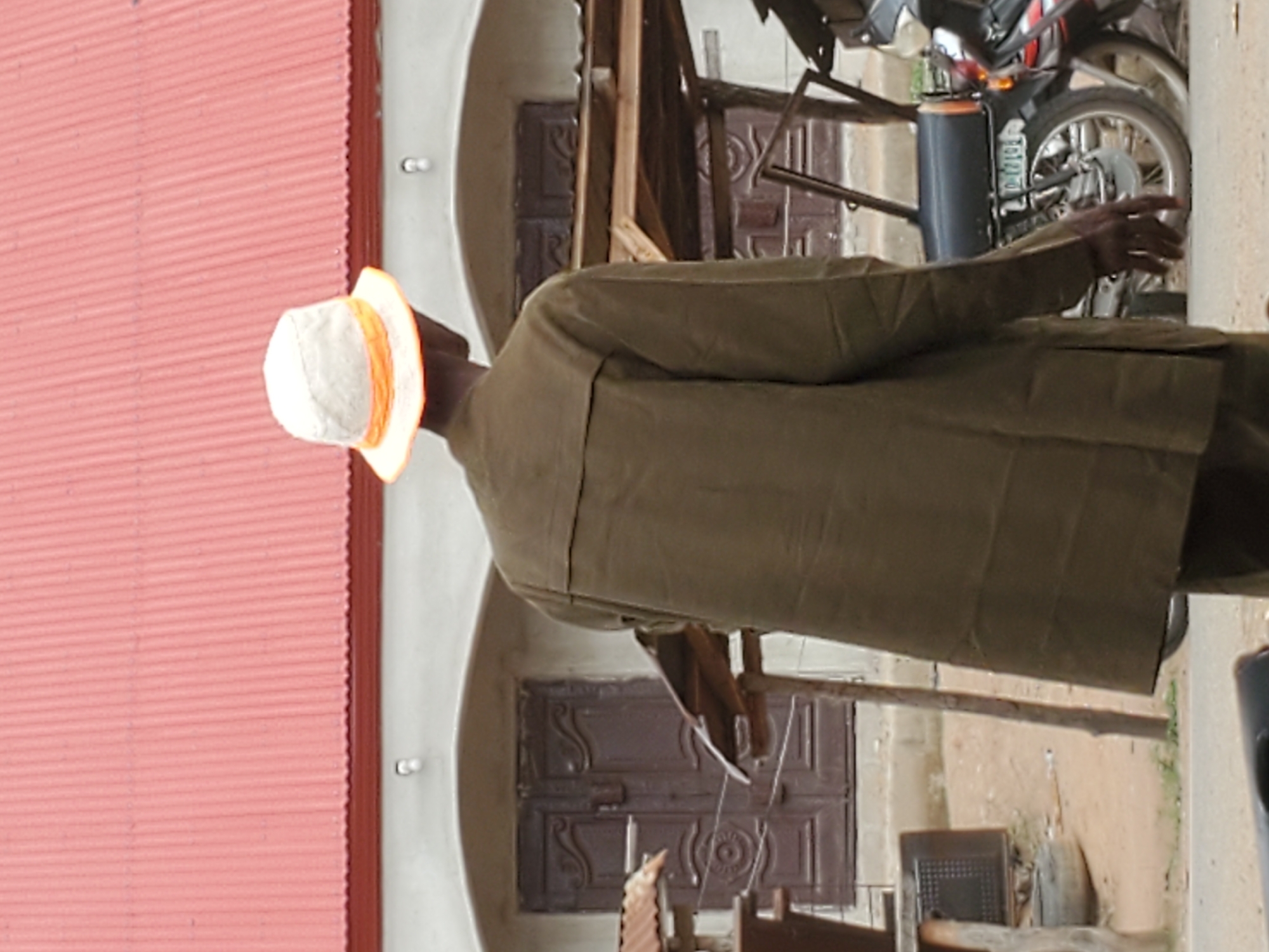 The image shows a man wearing a hat and a green coat, standing in front of a building with a red corrugated roof in Mararaba, Nigeria. He is reaching out towards a motorcycle. A double door with decorative carvings is visible behind the man and the motorcycle. A chair is also present in the background.