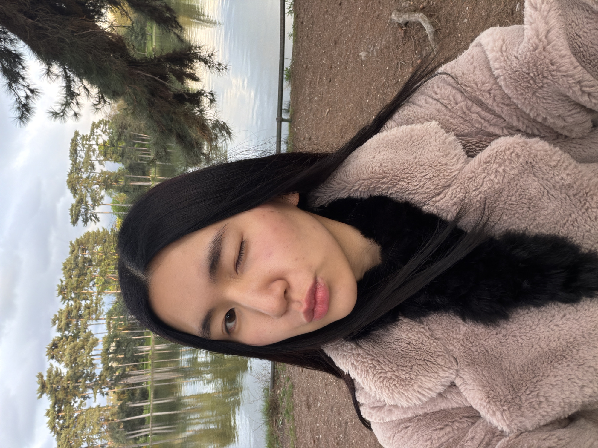 A woman is angled in a portrait-style shot, appearing outdoors near a body of water. The woman has black hair, one eye closed, and a neutral expression. She is wearing a tan-colored, furry jacket with a black fur collar. Behind her are trees, some reflected in the water. Based on the vegetation and overall atmosphere, it is plausible this photograph was taken in a park near a lake or pond, potentially in Paris, France.