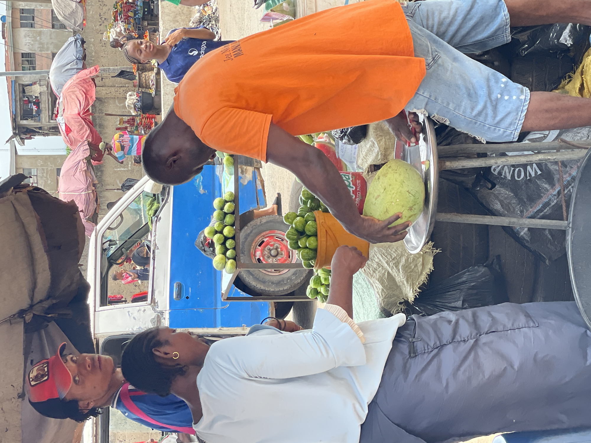 In the bustling city of Onitsha, Nigeria, a vibrant street scene unfolds. An individual wearing an orange t-shirt and light blue jean shorts stands behind a portable fruit stand. He's holding a large, light green pomelo and appears to be cutting it on a round metal tray with scissors. The fruit stand is a blue and white pick-up truck converted for commerce, loaded with what looks like small, green, unripe fruits and a container of tomato paste. The back of the truck holds additional supplies.
A woman in a white top and blue pants stands facing the fruit seller, apparently making a purchase. Another individual in a blue shirt is visible in the background, standing near a busy storefront or market area. The environment appears to be a lively marketplace with buildings, structures draped in colorful fabrics, and signs visible in the background. The day seems bright with ambient daylight.
