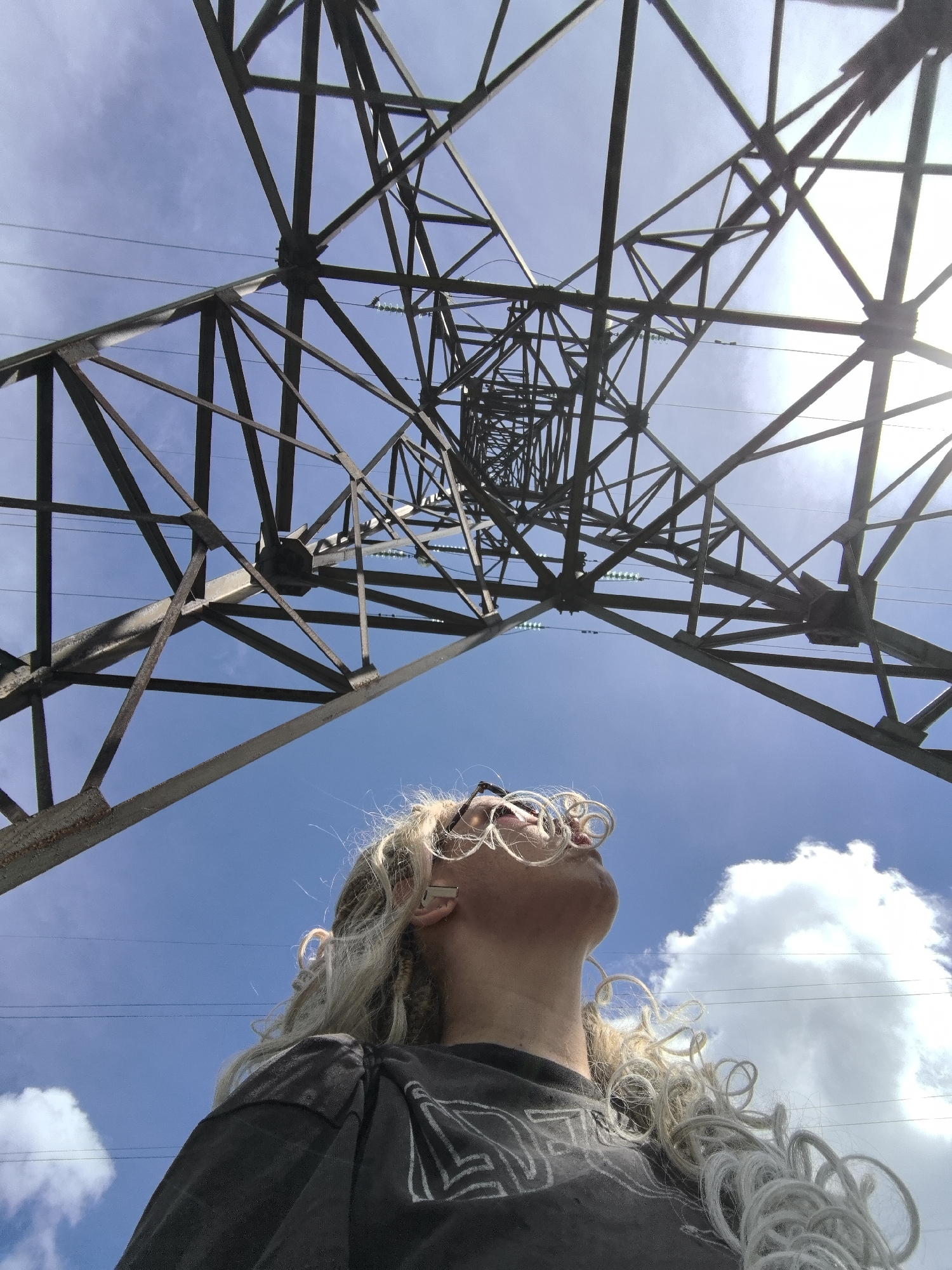 An upward-angle, medium shot captures a person with light blonde, curly hair and round glasses looking up at a large metal power line tower against a bright blue sky with scattered clouds. The person is wearing a dark t-shirt and is positioned in the lower right quadrant of the frame. The tower, made of dark metal beams forming a complex geometric structure, dominates the upper portion of the image, with power lines visible stretching across the sky. The sun is bright, creating a lens flare on the right side of the image. The image appears to be taken outdoors, possibly in an open area within the city of Noviy, Russia.