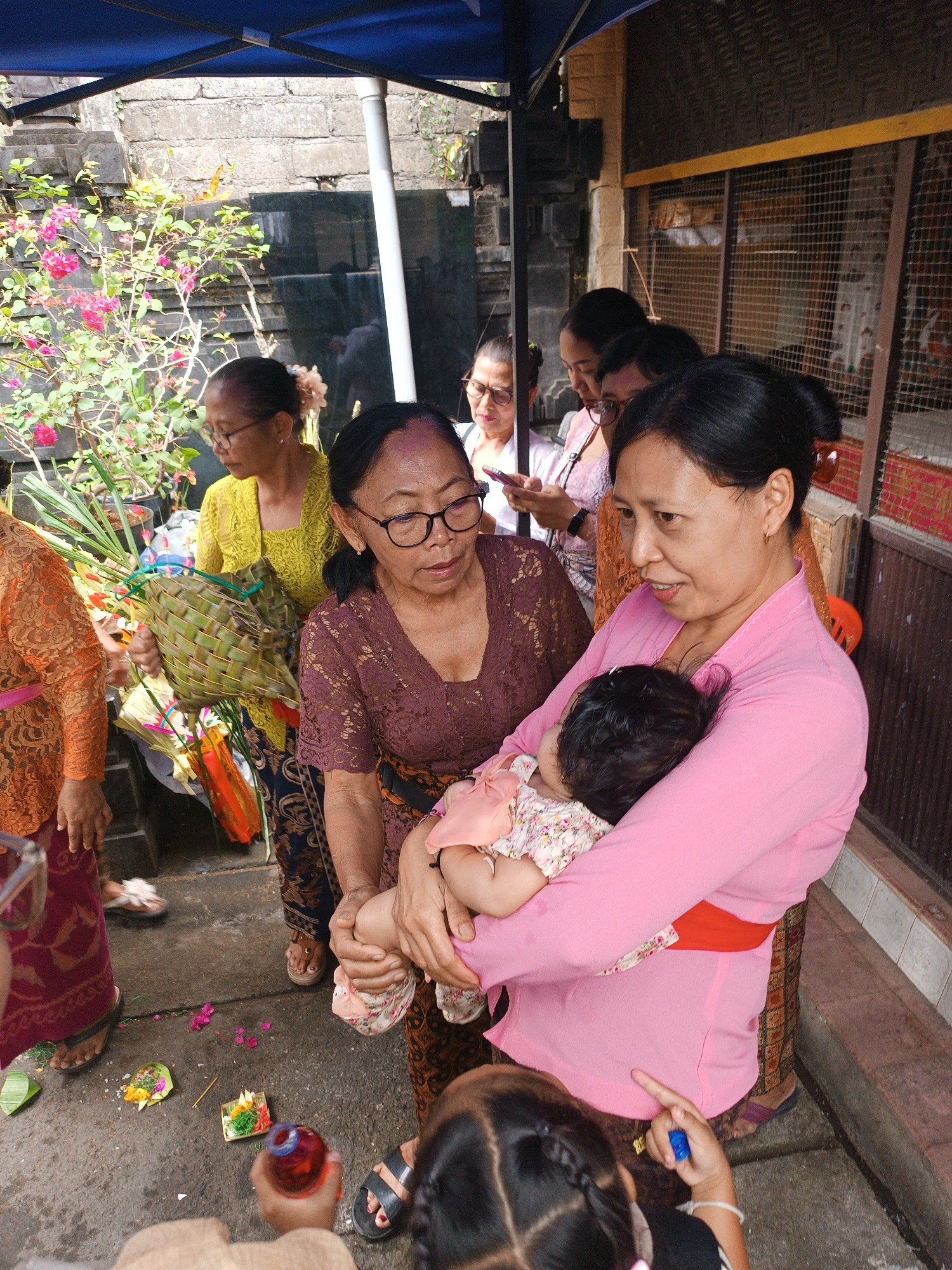 A group of women and a baby are gathered under a blue canopy. The women are wearing traditional Indonesian attire. One woman in a pink top is holding a baby. Another woman in a maroon lace top is looking down. A woman in a yellow top is holding a woven basket filled with greenery. The ground is paved with stone and has some offerings scattered on it. In the background, there is a stone wall and a caged structure. The scene appears to be outdoors, possibly at a temple or a ceremonial site in Tabanan, Indonesia.