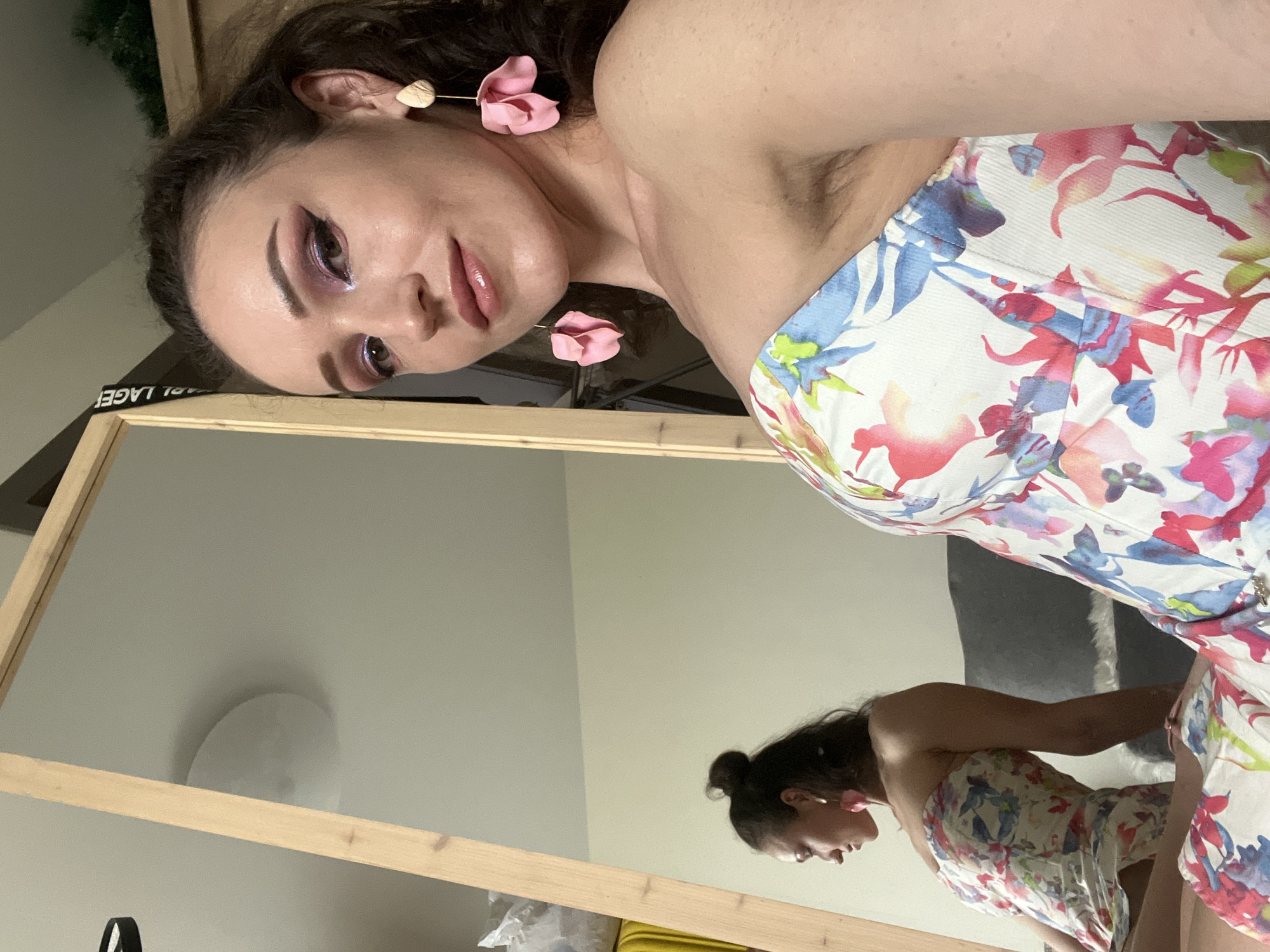 A young woman with dark hair and a floral strapless dress is posing in a room. She has on dramatic eye makeup and large pink flower earrings. Her reflection is visible in a large mirror, where she is seen from the side. The room appears to be a bedroom or dressing area, with a bed or sofa visible in the background. There are also some items on a shelf, including what appears to be a yellow suitcase. The overall impression is that of a selfie taken in a personal space.