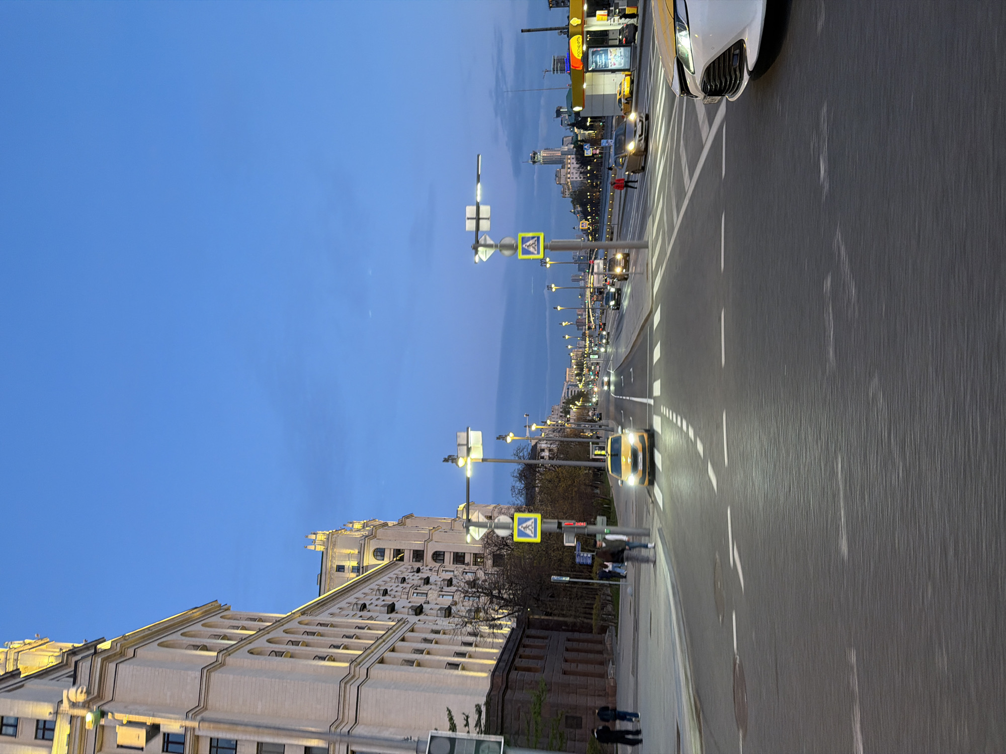 The image depicts a street scene in Moscow, Russia. A large, light-colored building stands on the left side of the image. The building features arched windows, decorative molding, and a tiered roof. A road occupies the right side of the image, showing multiple lanes with painted lines and illuminated streetlights. Cars are visible on the road, with headlights on, moving in both directions. Pedestrian crossings are marked with striped white lines, and a pedestrian crossing sign is visible with a yellow triangle. The sky is a gradient of blue, suggesting twilight or early evening. Pedestrians are seen walking on the sidewalk next to the building.