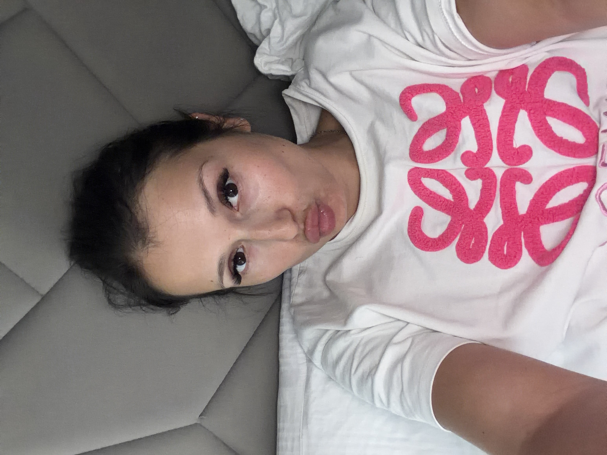 A woman is lying on a gray, quilted surface next to a white, rumpled sheet. She is wearing a white T-shirt with a pink design on the front. Her dark hair is pulled back, and she has brown eyes. She is looking directly at the camera with her lips slightly pursed. The photo appears to be taken from an elevated perspective.
There is no location information in the image.