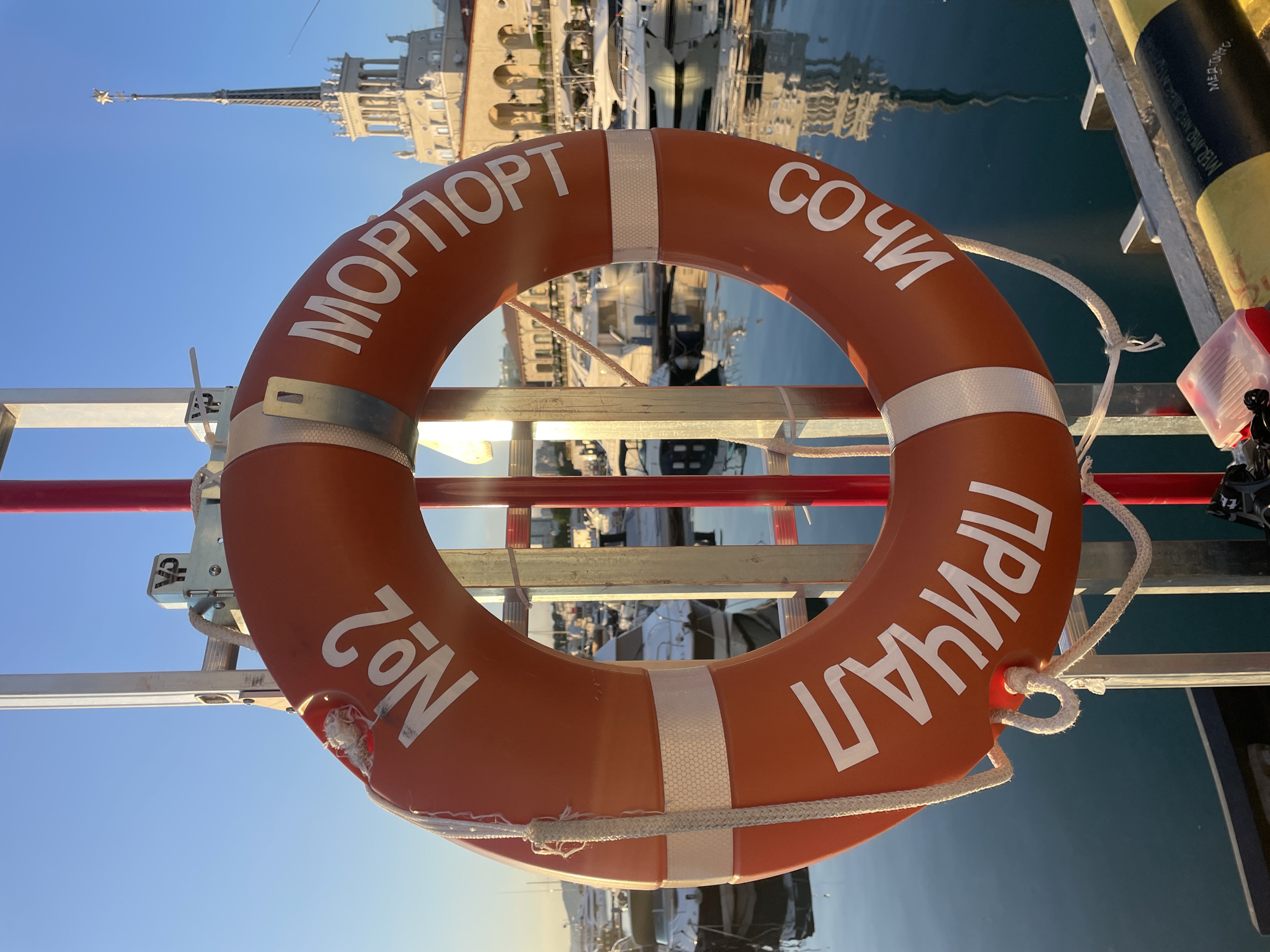 This image shows an orange life preserver hanging on a metal railing at a harbor. The life preserver has white text written on it in Cyrillic. The text reads "МОРПОРТ СОЧИ №2 ПРИЧАЛ" which translates to "Seaport Sochi Pier #2". The life preserver is positioned centrally in the frame.
In the background, a clear blue sky is visible, reflecting on the calm, dark blue water of the harbor. Several yachts and boats are docked, their reflections shimmering on the water's surface. A large, ornate building with a tall spire, characteristic of Soviet-era architecture, stands prominently in the distance, further suggesting a port or harbor setting. The lighting indicates it is daytime, likely late afternoon or early morning due to the warm glow on the building. The scene conveys a sense of tranquility and readiness, with the life preserver being a prominent safety object in this maritime environment. There are no people visible in the image.