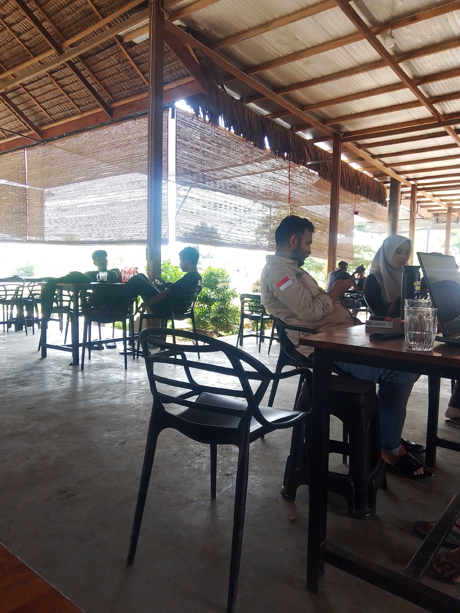 The image depicts an outdoor seating area at a cafe, possibly in Meunasahmee, Indonesia, with a thatched roof and bamboo blinds. In the foreground, a man wearing a tan shirt with an Indonesian flag patch on the sleeve is seated at a wooden table, looking down at his phone. Beside him, a woman in a hijab is looking at a laptop. A glass of water and a laptop are on the table. In the background, several other individuals are seated at tables, some appearing to be engaged in conversation. The environment is casual and relaxed, suggesting a typical cafe scene.