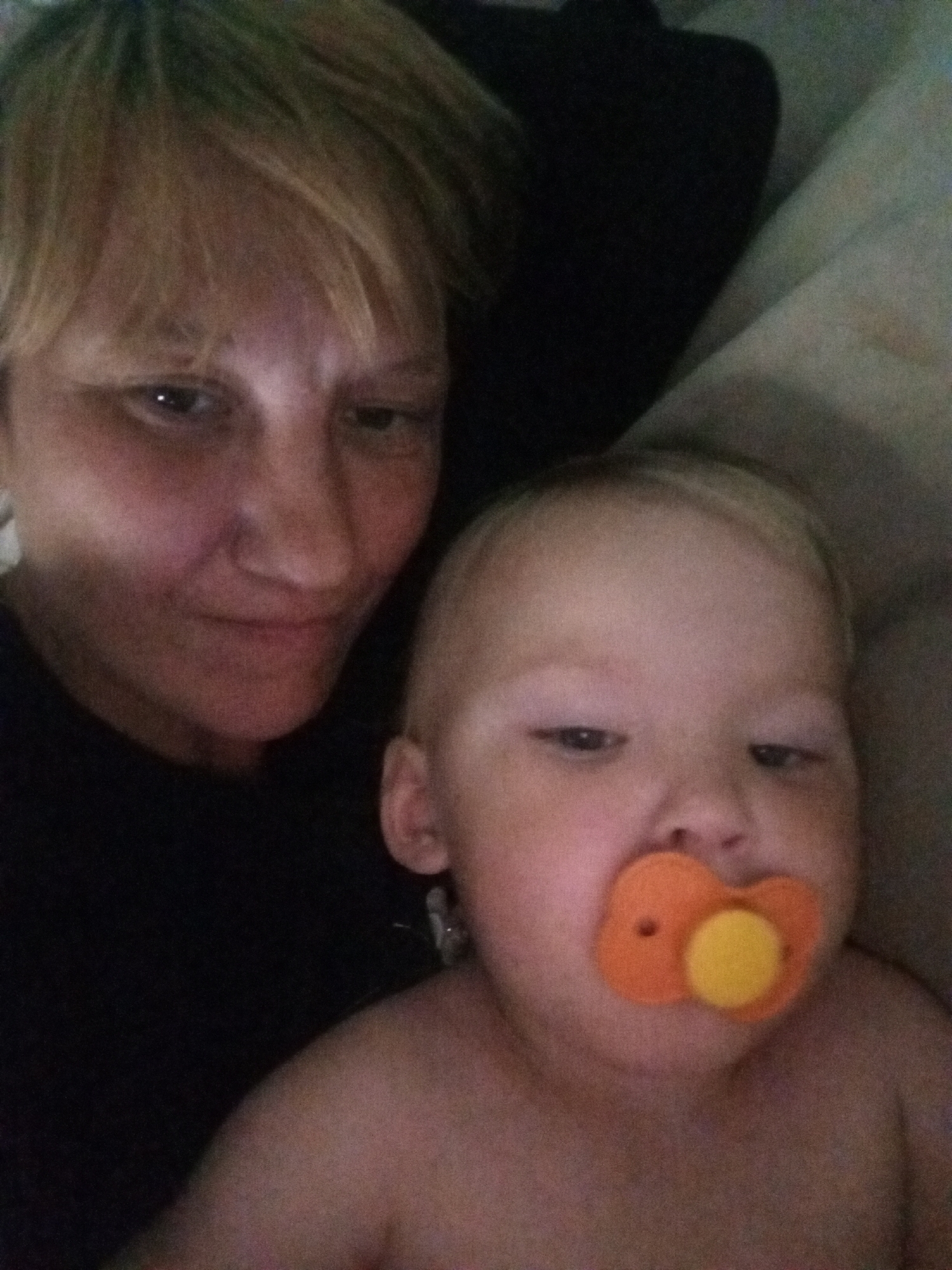 The image shows a selfie with two people, a woman and a baby, both appearing to be indoors, possibly in a domestic setting. The woman, in the foreground on the left, has blonde hair and is looking towards the camera. She is wearing a black top. The baby, positioned to the right and slightly behind the woman, has blonde hair and is wearing an orange pacifier. The background is dark and out of focus, with hints of fabric that suggest soft furnishings like bedding or a blanket. The scene is dimly lit. There is no indication of the city of Buraly, Russia, or any specific urban location within the image itself.
