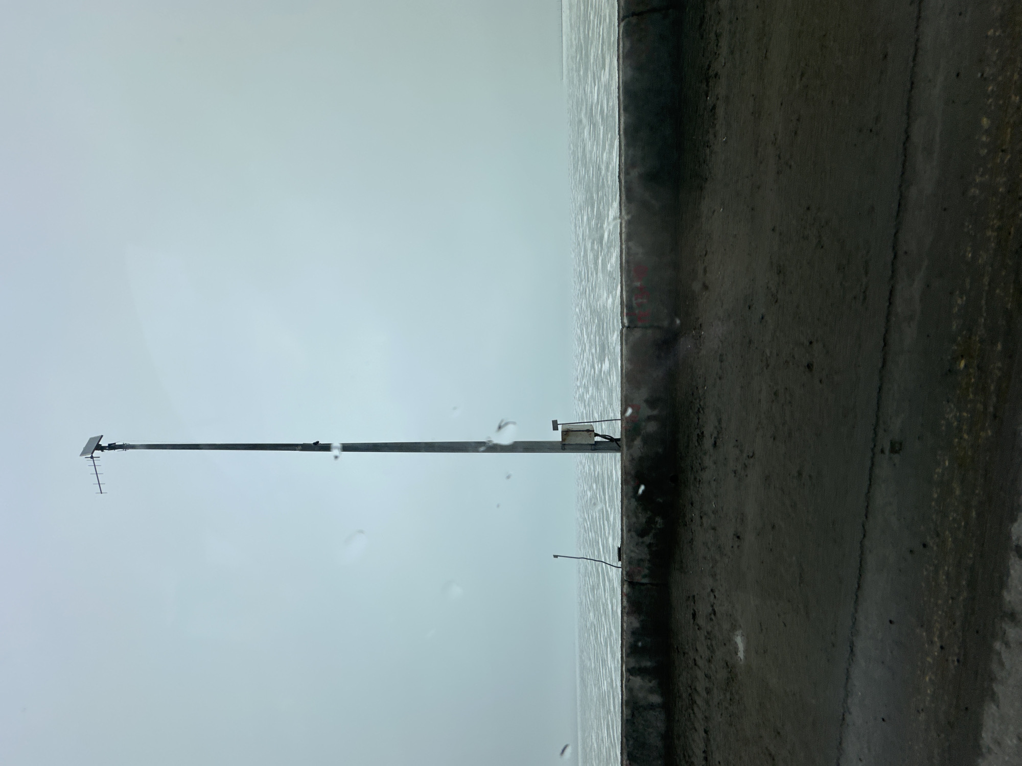 A vertical, long-shot image depicts a concrete wall and the sky/waterline in Chaikovsky, Russia. The dark concrete wall dominates the right portion of the frame. To the left of the wall, the horizon line separates the water from a cloud-covered sky. A tall, thin metal pole with antenna-like extensions rises vertically along the left side of the frame. Raindrops are visible on the lens or window from which the photo was taken.