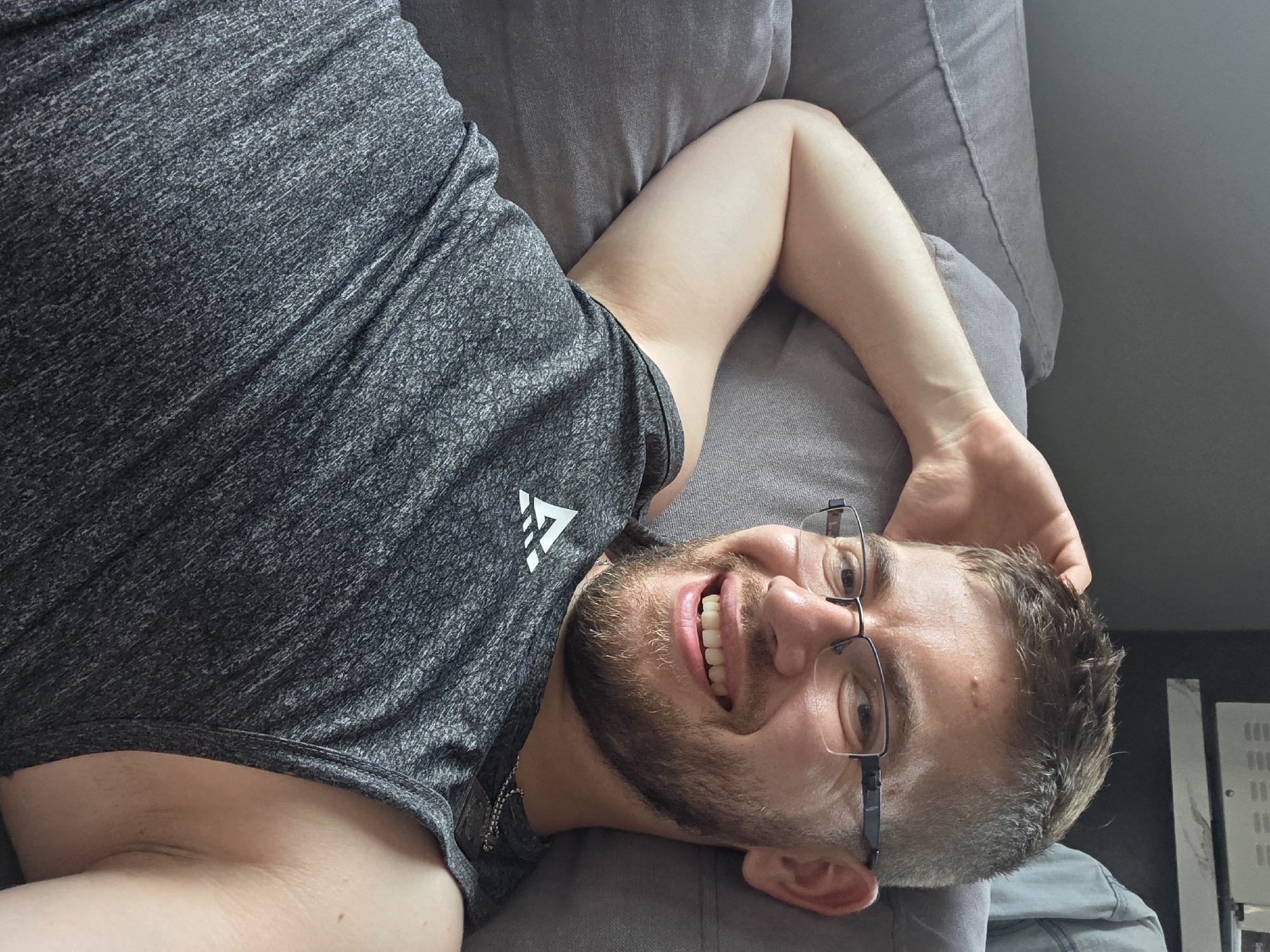 A man is lying on a gray couch, smiling. He is wearing a gray athletic shirt with a white logo on the left breast. He has short hair and a beard, and he is wearing glasses. The location appears to be indoors. There is no indication of the city of Yagodovo, Bulgaria.