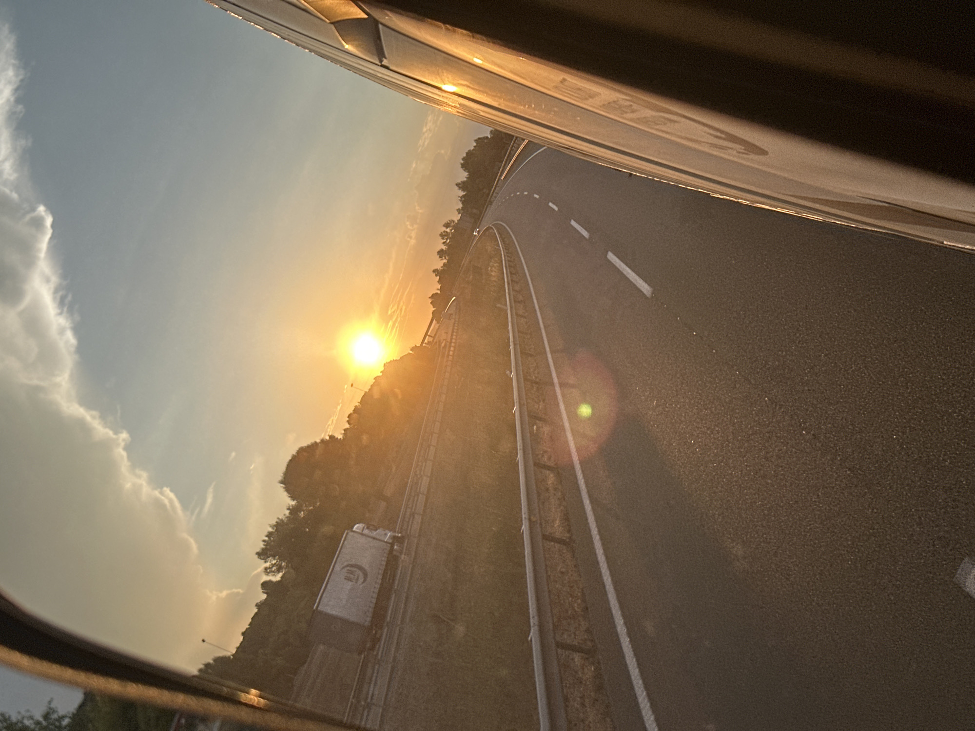 A rearview mirror reflection captures a sunset over a highway in El Montmell, Spain. The sun, a bright orb, casts a warm glow across a sky with scattered clouds. A semi-trailer truck is visible on the highway, traveling alongside a grassy embankment and a guardrail. The highway curves gently to the left, with white lines marking the lanes. The right side of the image is dominated by the reflection of the vehicle's side mirror, showing its metallic surface catching the sunset's light. Trees and hills form the background, silhouetted against the vibrant sky.