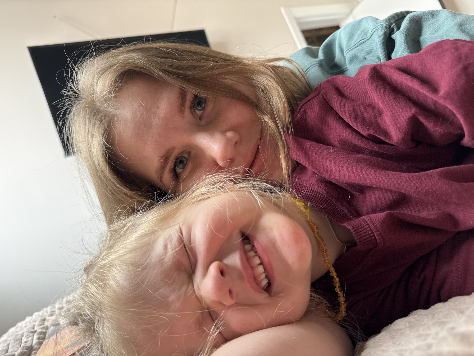 A close-up snapshot shows two individuals, a fair-skinned woman and a fair-skinned child, engaging in an intimate interaction. The woman has blonde hair and blue eyes, while the child also has blonde hair and is seen smiling widely, revealing their teeth. The woman is positioned above the child. The woman wears a burgundy sweater, while the child has a beaded necklace. The environment includes a TV visible in the background. The location is unknown, and there is no clear evidence of the location being Gornyi (Dvurechenskoe S/P), Russia.