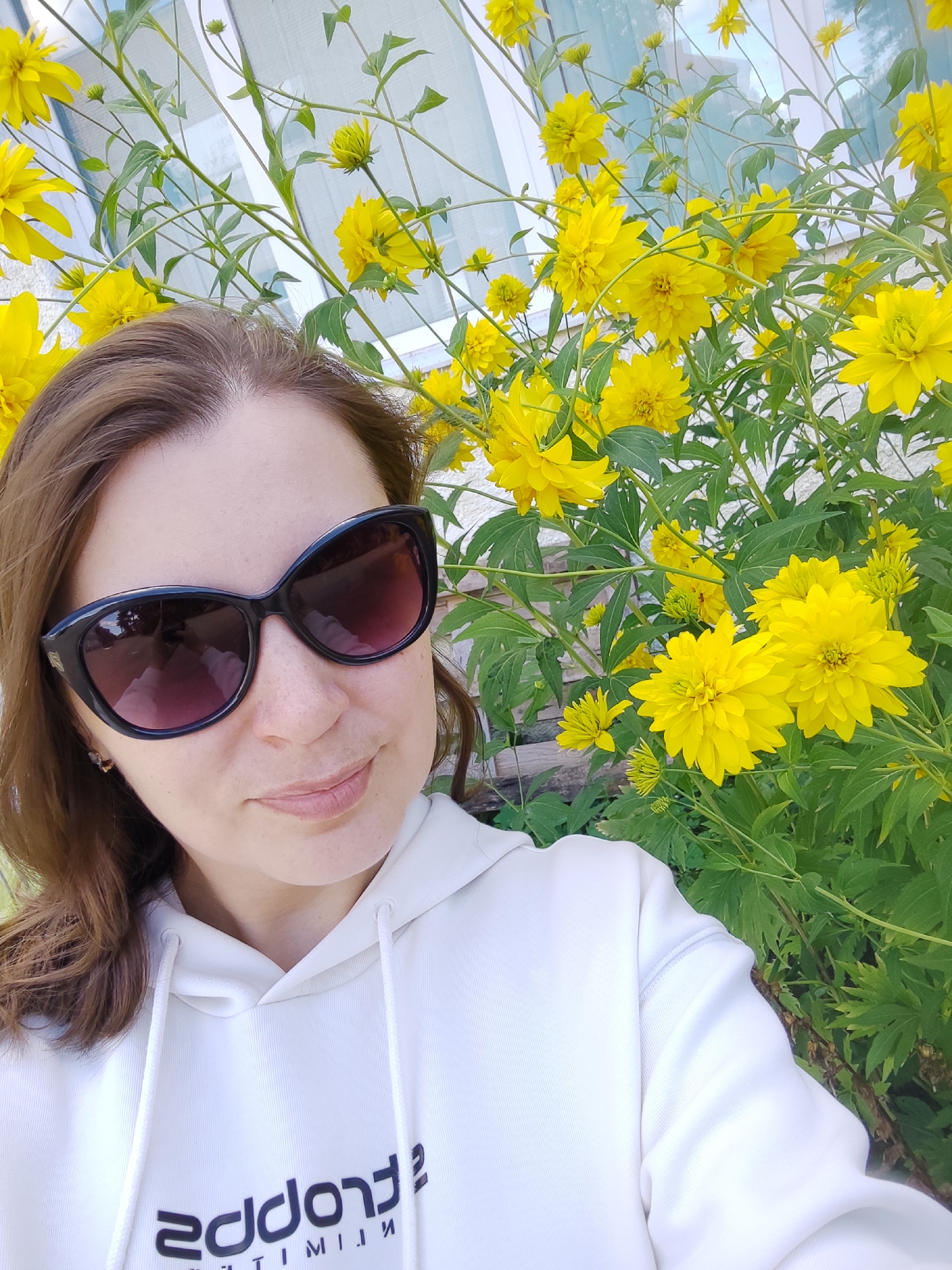 A selfie shows a woman wearing large black sunglasses and a white hoodie with black text. She is outdoors in front of a large bush of bright yellow flowers. The background is slightly blurred, with hints of a building with windows and some greenery. The lighting suggests it is daytime.