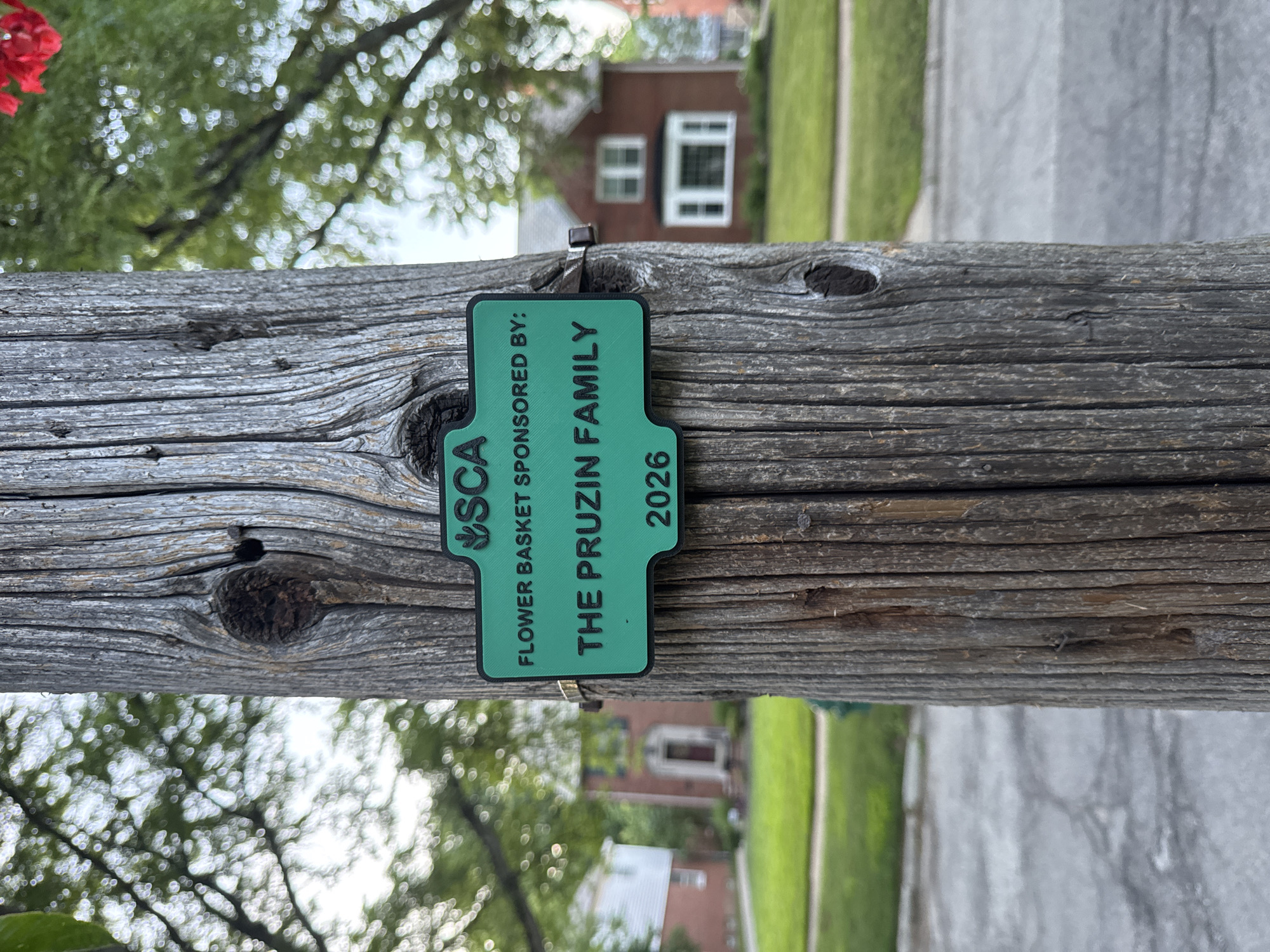 A green sign is attached to a weathered wooden utility pole in a residential area. The sign reads "SCA FLOWER BASKET SPONSORED BY: THE PRUZIN FAMILY 2026". In the background, out of focus, are houses and trees. The scene appears to be in Shaker Heights, Ohio, USA.