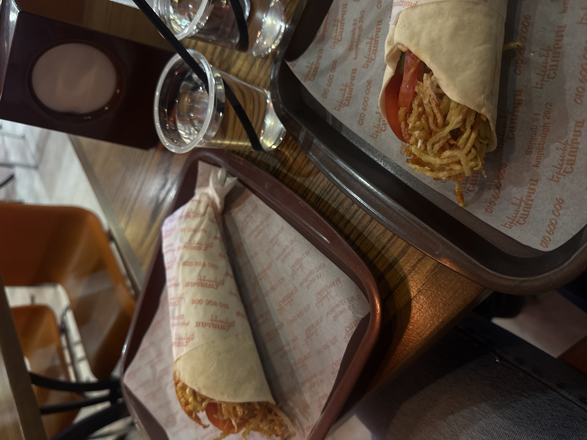 The image shows a tabletop setting with two trays of food and beverages, likely in a restaurant or cafe setting. Each tray holds a wrapped sandwich or shawarma, with visible fillings including what appears to be shredded potatoes and tomato. Next to the trays are two clear plastic cups with straws, possibly containing beverages like juice or soda. The restaurant/cafe is in Yerevan, Armenia, specifically at 91 Teryan st. and 29/2 Khorenatsi st., according to text on the wrapping paper. The environment seems casual, indicated by the wooden table and the chair visible in the background. A person wearing denim is visible.