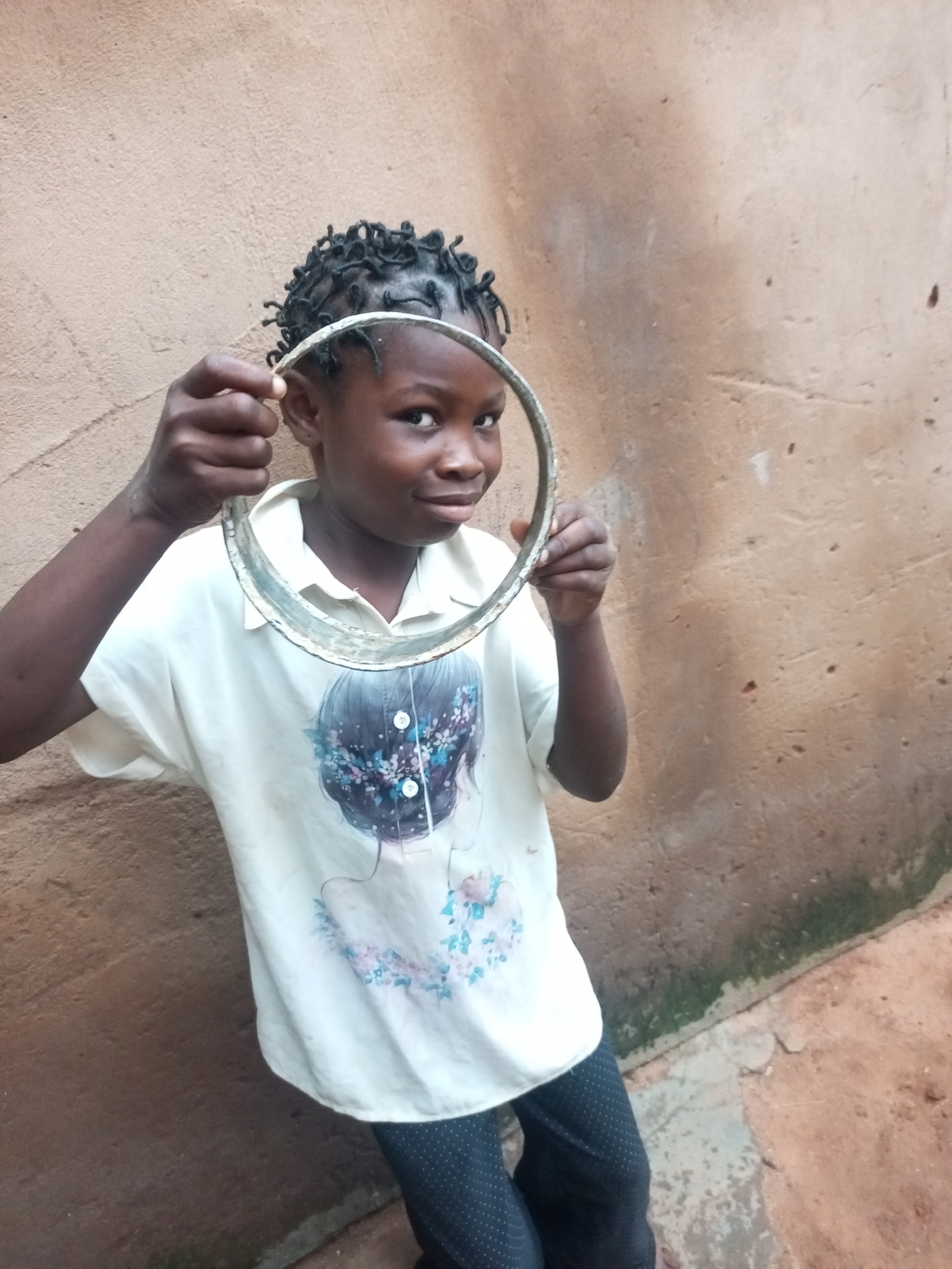 A young girl with dark skin and braided hair is standing in front of a textured, tan-colored wall, holding a metallic, circular object up to her face. She is wearing a white t-shirt with a printed image of a person's back with dark hair adorned with flowers, and dark pants with a white polka dot pattern. Her left arm is bent, and her fingers are wrapped around the circular object, which appears to be a metal ring or a small hoop. She is looking directly at the camera with a slight smile. The environment suggests an outdoor setting in a city, potentially Gombe, Nigeria, characterized by the earthy tones of the wall and ground, and a hint of green moss or algae along the base of the wall. The lighting indicates it is daytime.