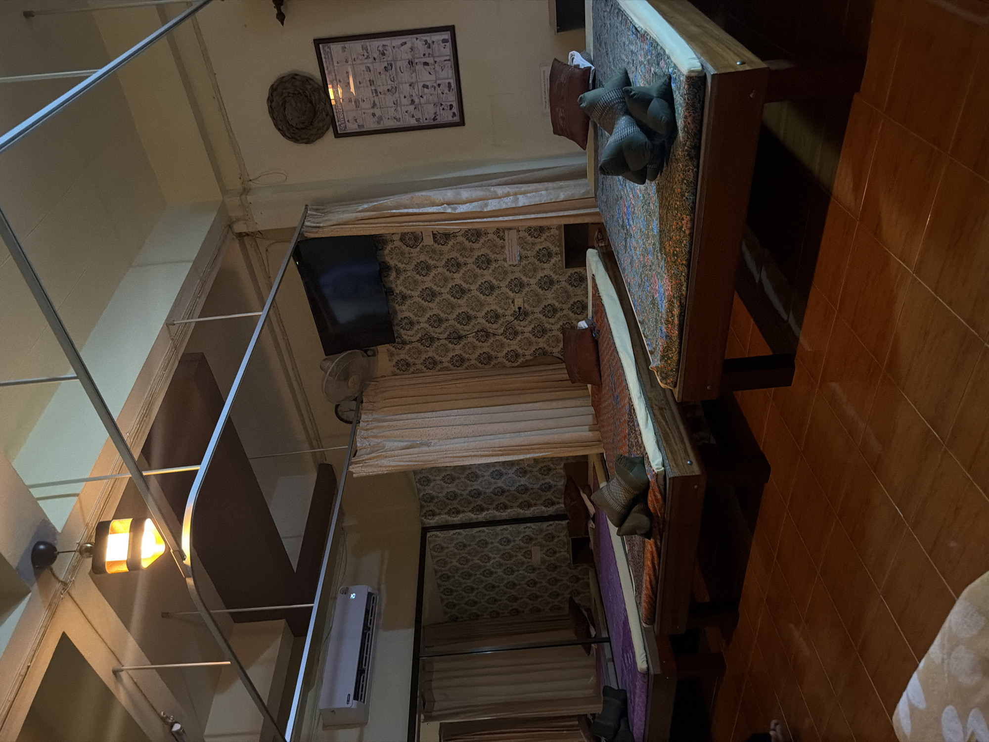 The image shows a room with three massage beds in what appears to be a spa or massage parlor in Baan Tai, Thailand. The room has a warm, ambient lighting, with a decorative lamp hanging from the ceiling. The beds are covered with patterned fabrics, and on each bed, there are pillows and folded towels. The walls are decorated with patterned wallpaper. On the right wall, there is a framed poster depicting various massage techniques. A television is mounted on the wall above a set of beige curtains. An air conditioning unit is visible on the left side of the room. Mirrors line the left wall, reflecting the massage beds and curtains. The floor is tiled with brown ceramic tiles. There are no people visible in the image.