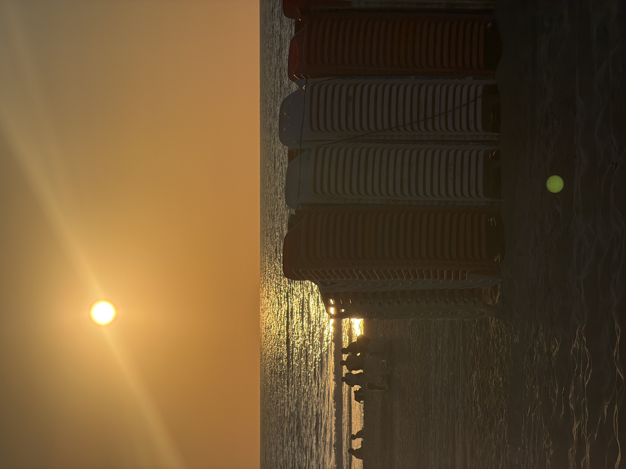 The image depicts a sunset at Bat Yam beach in Israel. The sun, a bright orb with visible rays, is positioned in the upper left quadrant of the sky, casting a warm orange glow across the scene. The sky is a gradient of orange and yellow, transitioning to a darker hue towards the horizon.
In the foreground, several stacks of beach chairs are neatly arranged, with alternating colors of orange and white visible. These chairs are the dominant feature in the right half of the image. The sand, a textured surface with visible ripples and footprints, fills the lower portion of the frame. A faint green light artifact is present on the sand in the lower right corner.
The middle ground features the calm sea, with gentle waves lapping the shore. The water reflects the sunlight, creating a shimmering path across its surface. Along the shoreline, silhouetted figures of people are visible. Some are seated, appearing to be gathered in small groups, while others may be standing or walking. A backpack is visible near one of the groups. These figures are indistinct due to the backlighting from the setting sun. The overall atmosphere is serene and tranquil, capturing a moment at the end of the day on the beach.