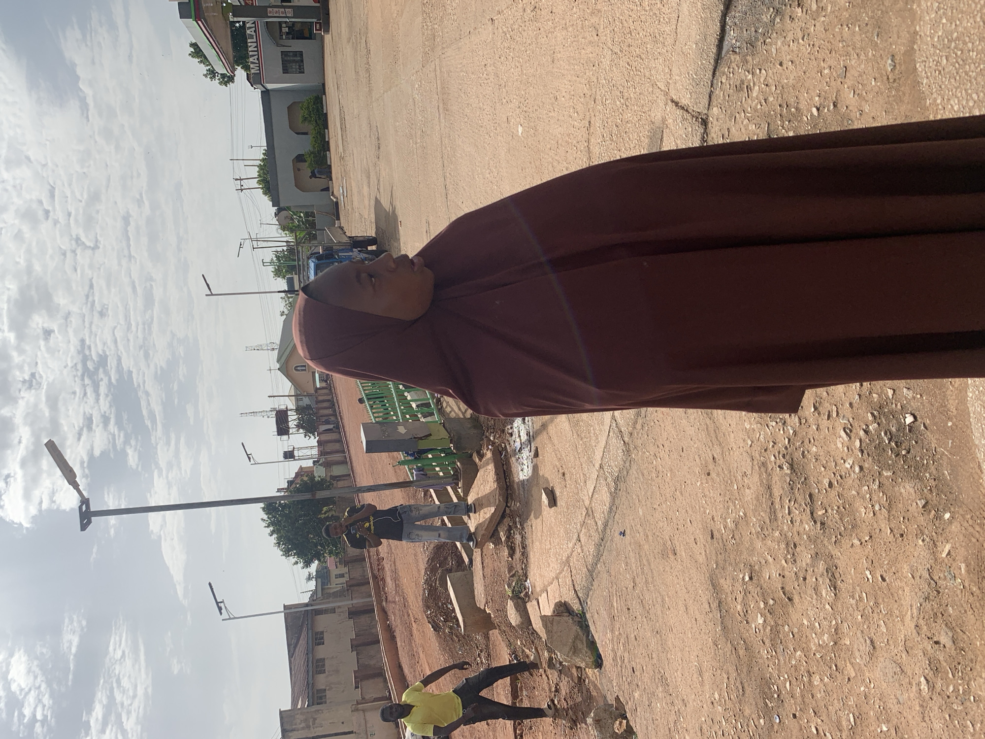 In Gura Top, Nigeria, this outdoor scene depicts a young woman wearing a maroon hijab in the foreground, with her profile facing right. Behind her, a man in a yellow shirt and dark pants stands on a dirt path, looking towards his left. Further in the background, another man in a dark t-shirt and light-colored jeans holds a smartphone. A blue truck is visible behind him. The environment is dusty and appears to be under construction or undergoing repairs, with piles of dirt and scattered rocks. A gas station with a sign reading "MAINLAND" is situated in the background, along with several buildings and streetlights. The sky is partly cloudy.