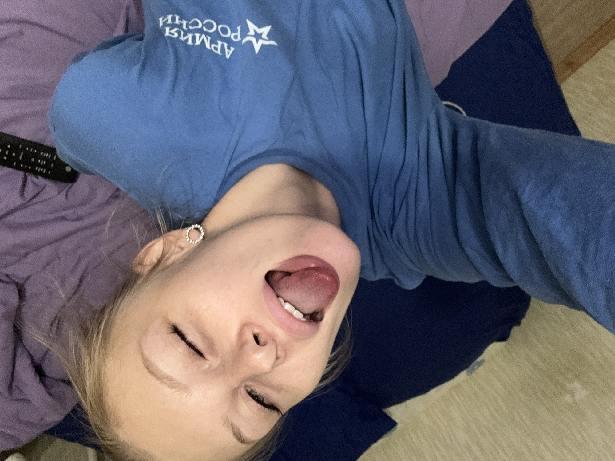 A young woman with blonde hair lies on a bed in what appears to be a bedroom. She is wearing a blue t-shirt with white text on it. Her eyes are closed and her tongue is sticking out. A remote control is visible on the purple blanket behind her head. The lighting suggests it is daytime. The location is described as the city of Kondratovo, Russia.