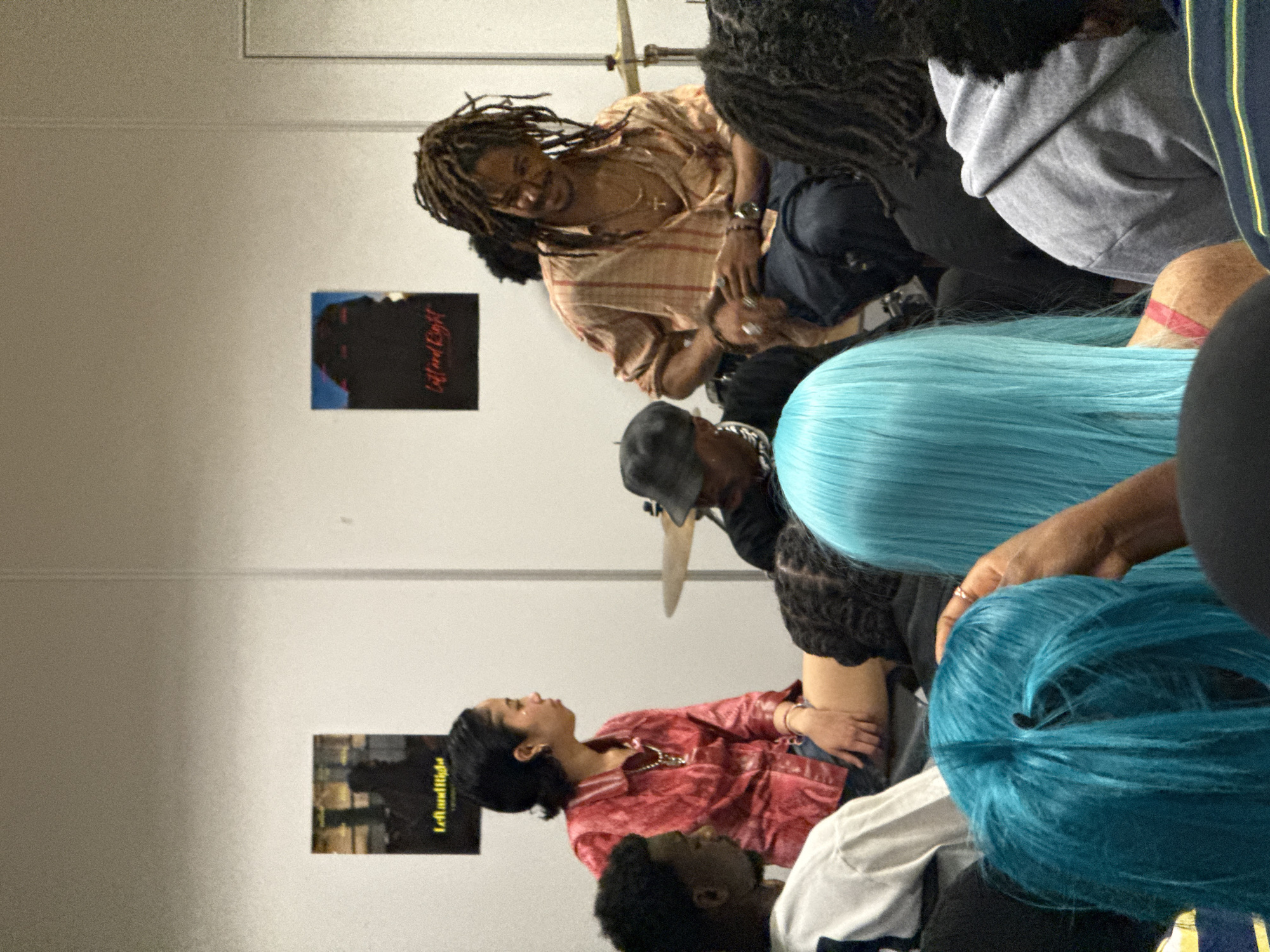 A group of people are gathered in a room, with one individual wearing dreadlocks seated on an elevated surface. Several attendees have their backs to the camera. There are two posters on the wall. The setting appears to be indoors. The image does not contain enough detail to specify a location in Saint-Ouen-sur-Seine, France.