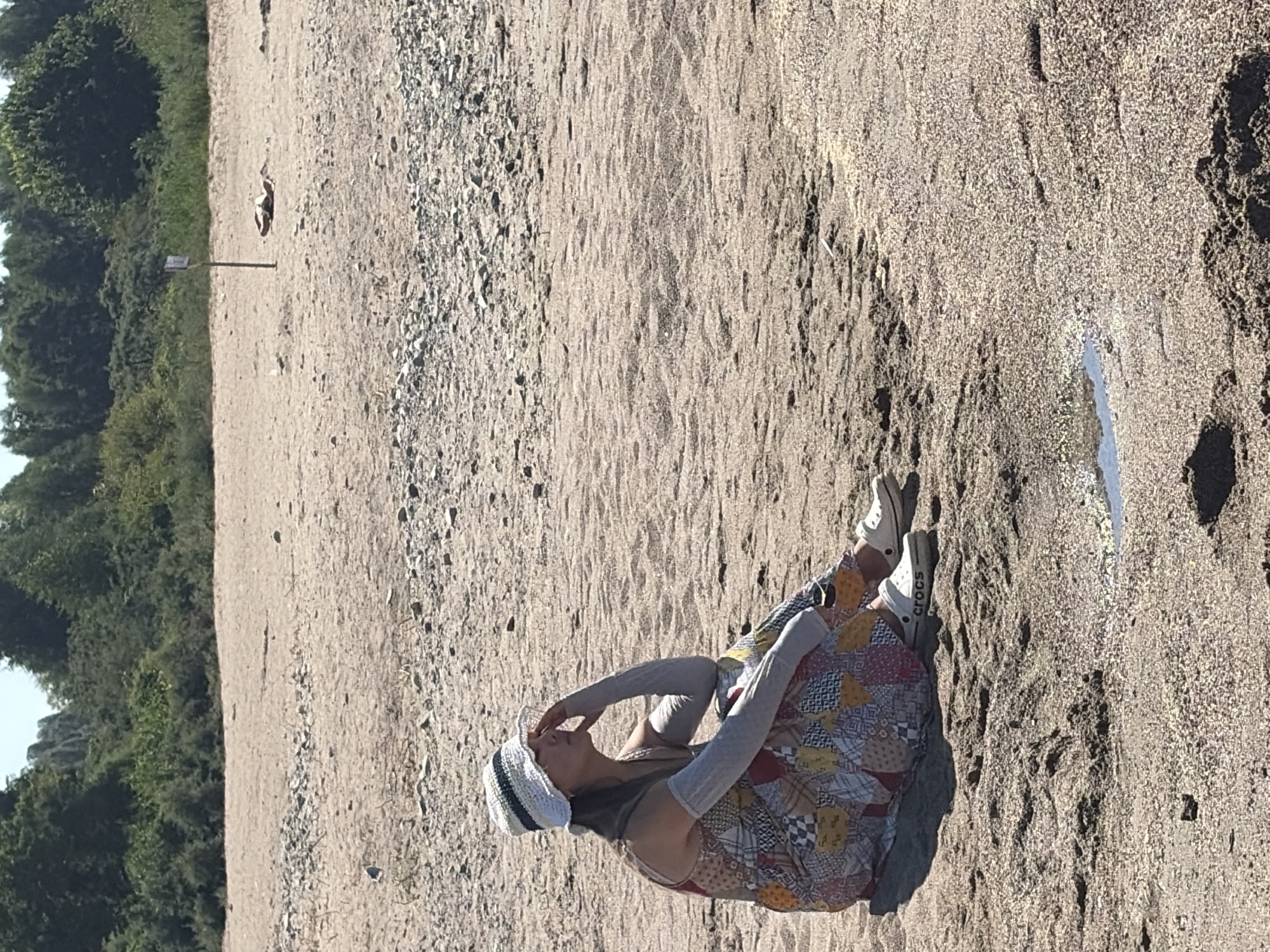 A woman is sitting on a sandy beach, facing right, with her left hand on her forehead and her eyes closed. She is wearing a patchwork dress, a white sun hat, and gray arm sleeves. Her feet are clad in white Crocs. The beach is covered in sand and small pebbles, with a small puddle of water in the foreground. In the background, there is a line of trees and a signpost with a person lying down near it.