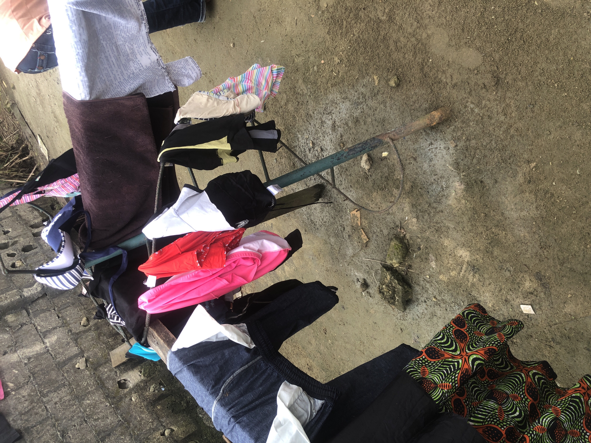The image depicts a clothesline with various garments drying outdoors in what appears to be a residential area. The clothesline is supported by a green metal stand, and a rough concrete wall forms the backdrop. Several pieces of clothing are visible, including what look like jeans, a pink garment, a red sequined top, a white shirt, a dark-colored top, and a striped garment. The ground is a dirt or sandy surface, with some rocks and debris scattered around. The scene suggests a typical domestic activity of laundry being air-dried. There are no people visible in the frame, but the presence of laundry and the clothesline indicates recent human activity. The overall impression is one of a humble outdoor setting, possibly in a less developed urban or semi-urban area.