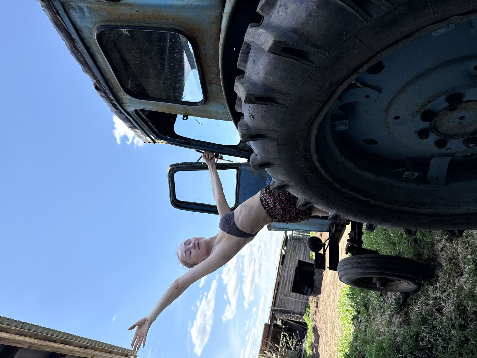 A low-angle shot of a female figure suspended from the door frame of a blue tractor in Nizhnyaya Chermoda, Russia. The woman, wearing a dark bra top and floral shorts, is hanging from the tractor's open door. Her body stretches diagonally across the frame, with her left arm reaching toward the lower left corner. The tractor, displaying rust, dominates the upper portion of the scene, with a large tire visible. A derelict wooden structure stands in the background, set against a blue sky. Vegetation and soil are evident in the lower right quadrant.