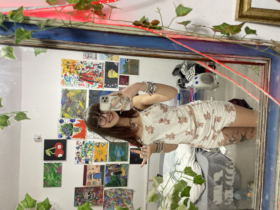 A person in a teddy bear pajama set poses in front of a mirror. A variety of art pieces are displayed on the wall behind them, alongside a small space heater. The mirror is surrounded by leafy vines and a red light strip. The person is wearing glasses, bracelets, and rings and holding up their phone to take a photo.