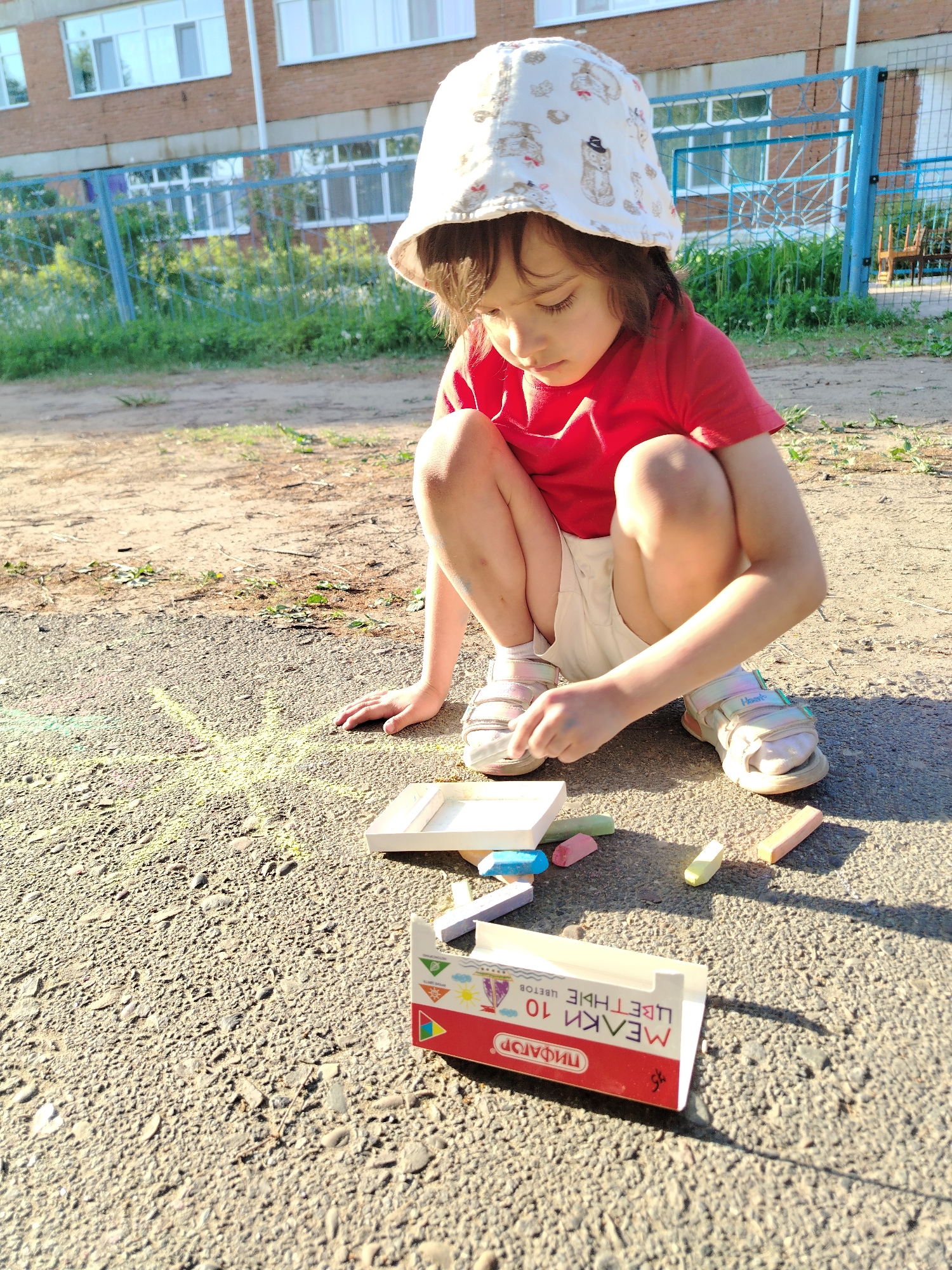 A young child wearing a white bucket hat, red t-shirt, and light shorts is squatting on a paved surface, possibly a sidewalk or driveway. The child is looking down at a box of colored chalk and some loose chalk sticks scattered nearby. A drawing in yellow chalk is visible on the pavement. In the background, there's a brick building with white-framed windows, a blue metal fence, and some greenery. The location is Noviy, Russia.
