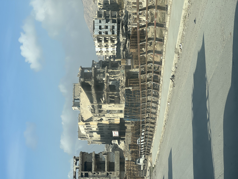 Buildings stand in ruins, skeletal structures against a light blue sky with scattered clouds. Debris is visible, with wooden pallets stacked high along the roadside. A gray asphalt road stretches along the right side, marked with shadows and the visible interior of a vehicle. Distant mountains can be seen in the background, under the hazy skyline.