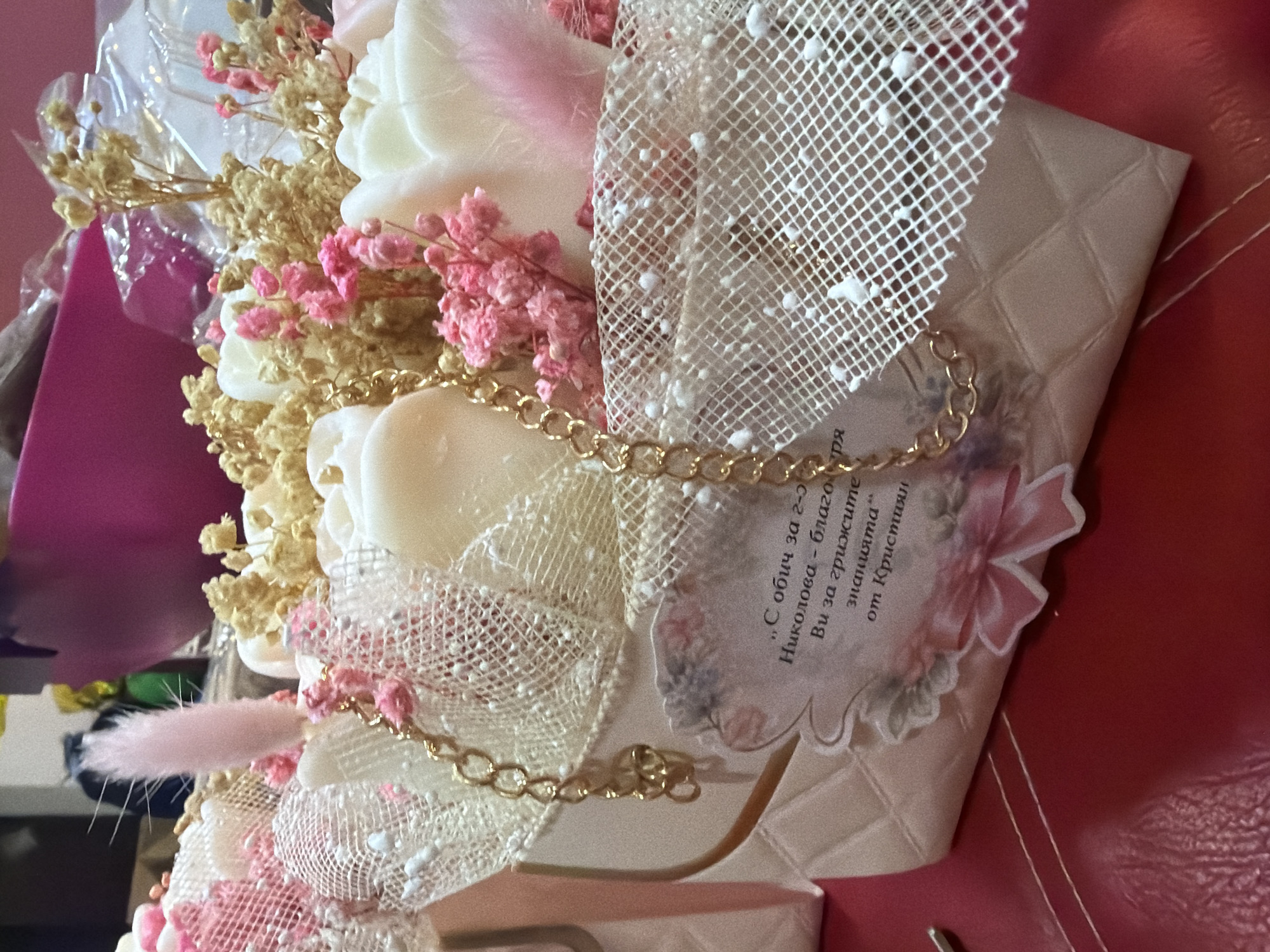 The image displays a close-up of a decorative arrangement, likely a gift, featuring white and pink artificial flowers or soap roses, interspersed with dried beige and pink floral elements. A white mesh-like fabric with textured white dots and a golden chain are draped across the arrangement, adding intricate detail. Resting on a beige, quilted-patterned surface, which appears to be a box or gift package, is a small decorative card.
The card contains text in Bulgarian, which translates to:
"With love for Mrs.
Nikolova - thank you
for your care
and knowledge"
from Kristiyan
The arrangement is set against a vibrant red background, possibly a table or fabric, with a blurry purple object partially visible in the upper left background. The overall impression is that of a thoughtful and elegantly presented gift, conveying appreciation from Kristiyan to Mrs. Nikolova. No activity is taking place, and the time of day or specific location is not discernible.