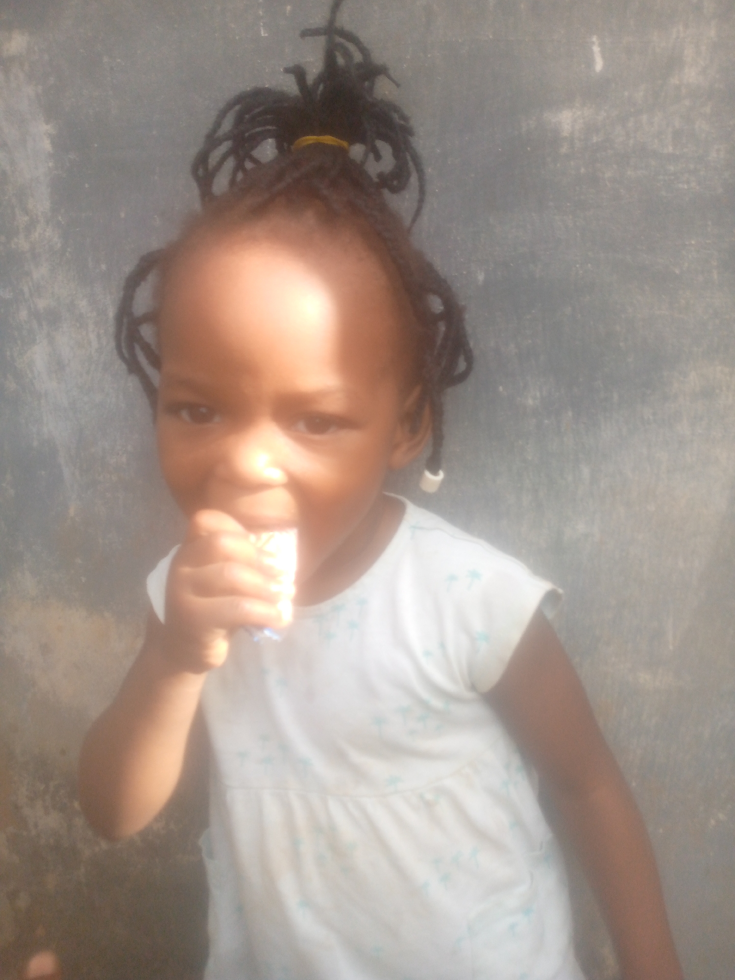 A young child with dark skin, wearing a white dress with a blue palm tree pattern, is holding a small, wrapped rectangular object in their hand and appears to be eating it. The child has dark hair styled in dreadlocks with a yellow hair tie. The background is a gray, textured wall with patches of discoloration. The location is the city of Unknown, Unknown.