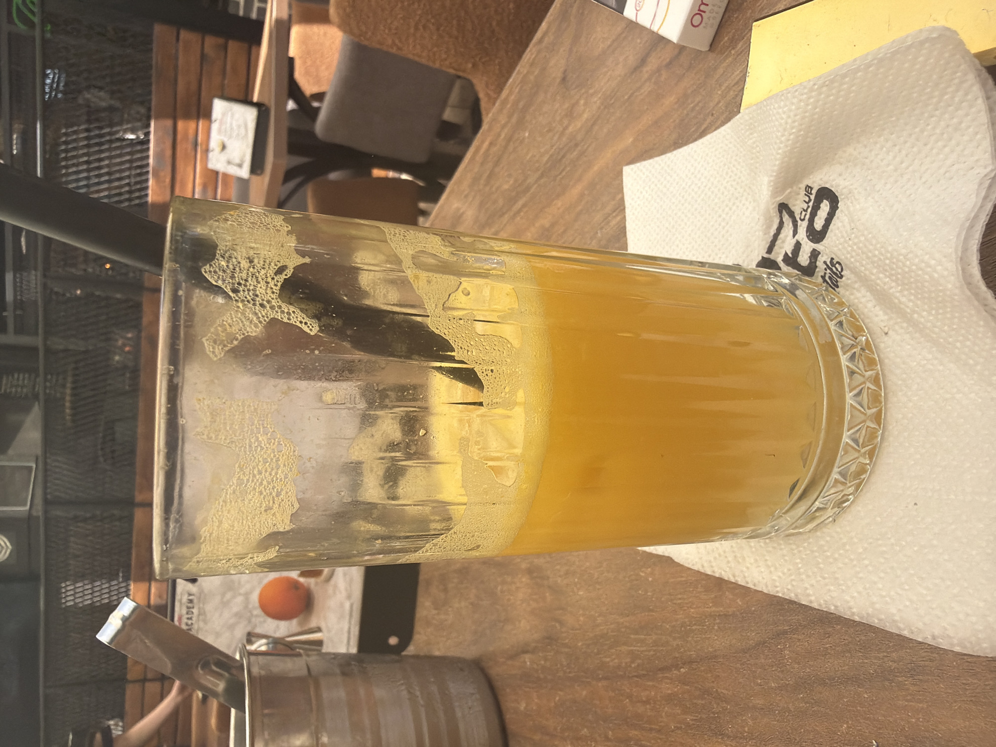 This image captures a close-up of a table setting at an establishment located in Shumen, Bulgaria. A tall, clear glass, approximately half-full with a yellow-orange beverage containing ice cubes, occupies the central foreground. A black drinking straw is inserted into the liquid. The upper interior walls of the glass exhibit dried, textured residue, consistent with pulp and foam. The glass rests on a white, patterned paper napkin featuring a partially visible black logo that includes the text "LEO CLUB" and "tails". To the immediate right of the napkin, a white and orange cigarette pack, partially obscured, and a yellow rectangular object are visible. The table surface is made of wood with a distinct grain pattern. To the left of the glass, a silver-colored metal bucket contains a metal utensil, possibly tongs. In the background, a person's arm is faintly visible near the bucket. Further back, a signboard displaying "ACADEMY" and a whole orange is present. The environment includes dark metal lattice work, wooden paneling or seating, and additional tables and chairs in muted tones, suggesting an indoor or semi-outdoor cafe or restaurant setting. No specific actions or interactions are discernible, and only a partial human presence is indicated.