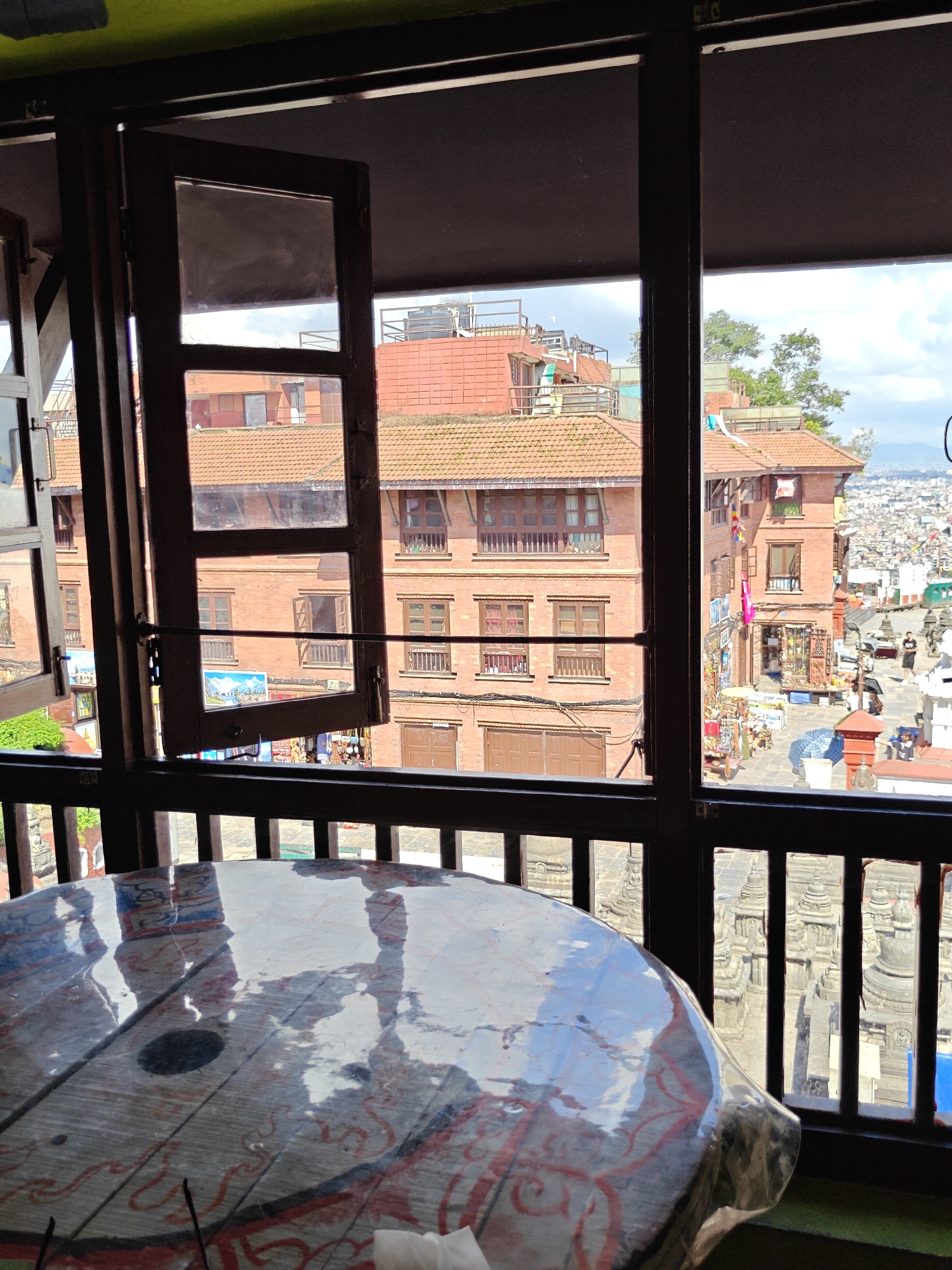 The image captures a view from a window, looking out onto a bustling street scene in Kathmandu, Nepal. In the foreground, a round wooden table covered with a reflective plastic sheet is visible. The table has a colorful, possibly painted, design. Beyond the table, a wooden window frame with an open casement is prominent, partially obscuring the view. Through the window, traditional brick buildings with tiled roofs are seen, characteristic of Kathmandu's architecture. The street below appears to be a marketplace, with several stalls and shops displaying goods. People can be seen walking around, and some are likely vendors. The background reveals a densely populated urban landscape with more buildings stretching into the distance, under a cloudy sky. The overall impression is of a vibrant, historical city environment.