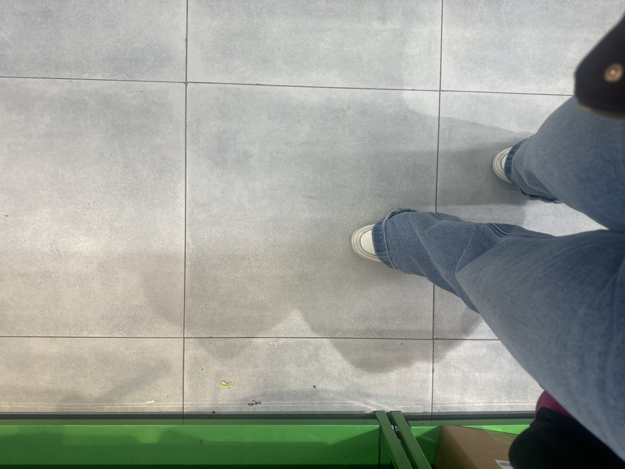 A first-person, high-angle shot captures a person standing on gray tile flooring. The person is wearing light-wash jeans and white sneakers. The lower portion of what appears to be a green shopping cart is visible along the bottom edge of the frame. The scene suggests an indoor environment. There is no information to suggest the location is Megion, Russia.