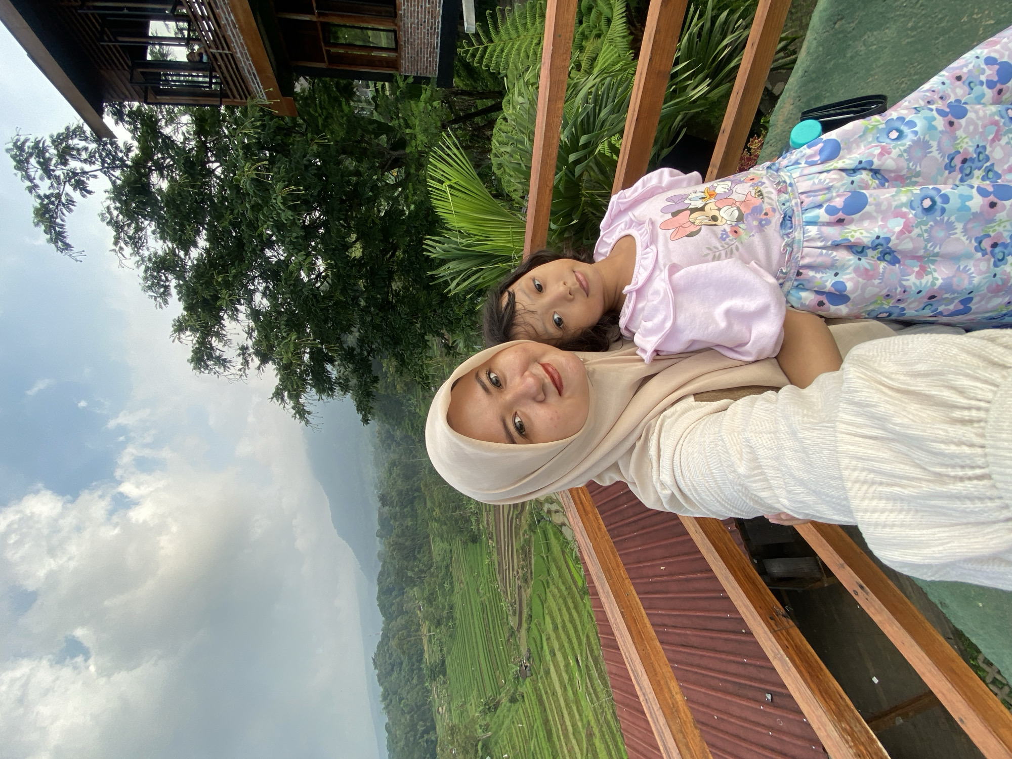 Two individuals are visible in the image. A woman wearing a hijab is at the forefront, likely holding the camera. A younger female, possibly a child, is positioned slightly behind her, wearing a light pink top and a dress with a floral pattern.

The setting appears to be a vantage point overlooking a rural landscape, likely the rice terraces of Selotapak, Indonesia. Terraced fields are visible in the background. A wooden railing suggests a balcony or viewing platform. The sky is overcast. A building is visible in the background.
