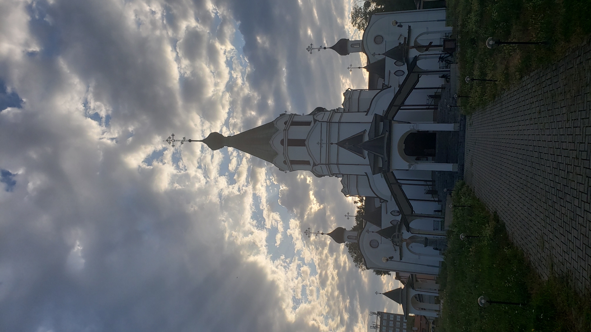 The image depicts an Orthodox church in Prešov, Slovakia. The church is a white, multi-domed structure with a prominent central spire topped by a cross. The sky above is dramatic, filled with dark, textured clouds with patches of sunlight breaking through. In the foreground, a paved walkway leads towards the church entrance, flanked by a grassy slope with small lampposts. The scene is set during what appears to be late afternoon or early evening, given the lighting conditions. There are no visible people engaged in any specific actions. The overall atmosphere is serene and somewhat solemn.