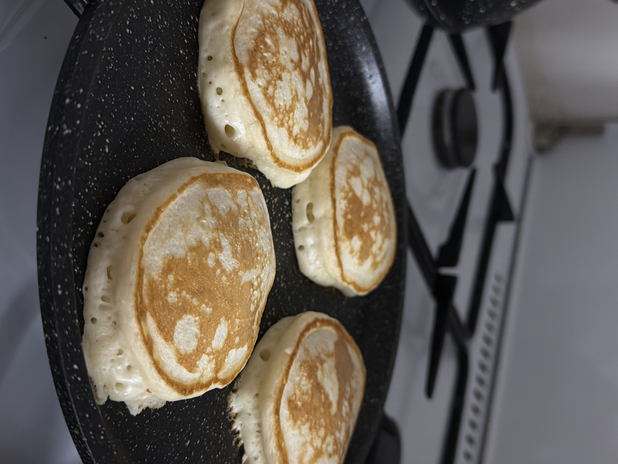 The image shows four pancakes cooking in a black frying pan on a gas stovetop. The pancakes are golden brown on top with visible bubbles. The stovetop is white with black grates. The image provides no information regarding location.