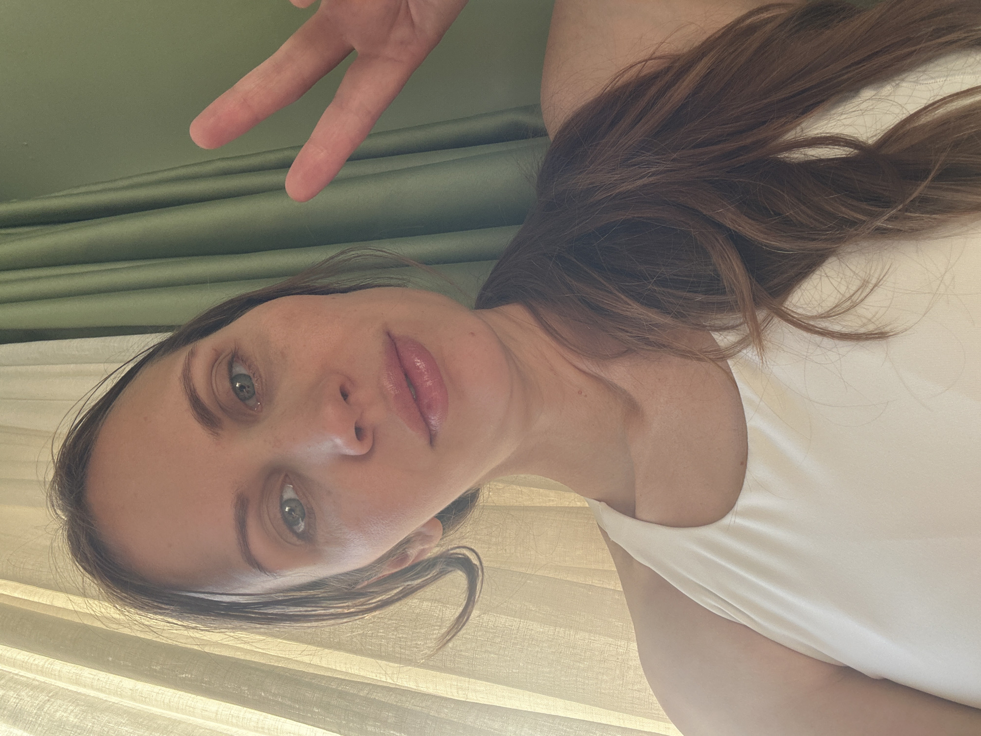This rotated image captures a close-up, casual portrait of a woman, likely a selfie, taken in what appears to be Fethiye, Turkey. The woman's head is positioned towards the left of the frame.
The main subject is a woman with long, wavy brown hair, which flows over her shoulders. She has light-colored eyes, possibly green or hazel, and a serene, direct gaze towards the camera. Her lips are slightly parted, revealing a natural sheen. She has a clear complexion and is wearing a simple, light-colored (off-white or cream) sleeveless top.
Her right hand is raised in the upper right corner of the image, making a clear "peace sign" (V-sign) with two fingers extended.
The background features two layers of curtains, suggesting an indoor setting with natural light. The upper part of the background shows a darker, olive-green pleated curtain. Below this, and behind the woman's head and upper torso, is a lighter, translucent curtain, likely sheer or a blind, which diffuses the daylight entering the room. The soft lighting indicates it is daytime.
No text is visible in the image. The overall impression is one of naturalness and calm.
