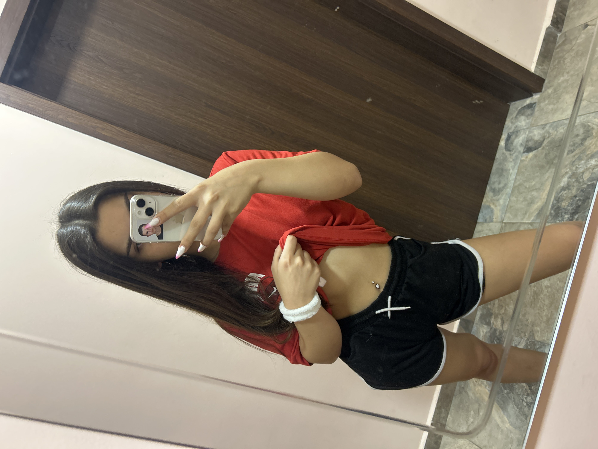 A young woman is taking a mirror selfie. She is wearing a red t-shirt and black shorts. Her hair is long and dark. She has a belly button piercing. The background is a room with a mirror and a wooden door. The location is not specified.