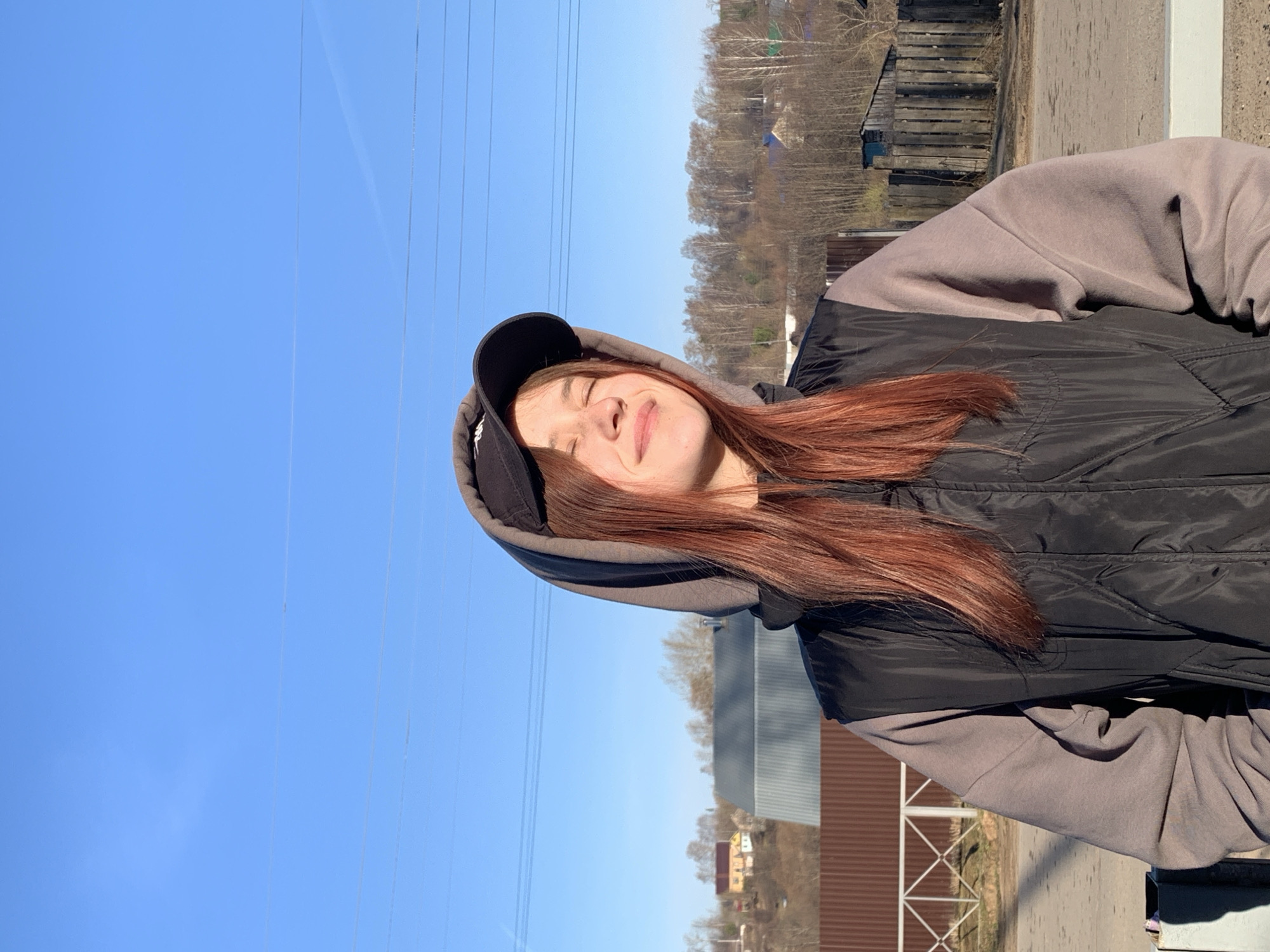 A person with long, reddish-brown hair is wearing a black baseball cap and a black and gray hoodie in Фоки, Russia. They are standing outside on a sunny day with a clear blue sky. In the background, there are power lines, trees, and buildings.