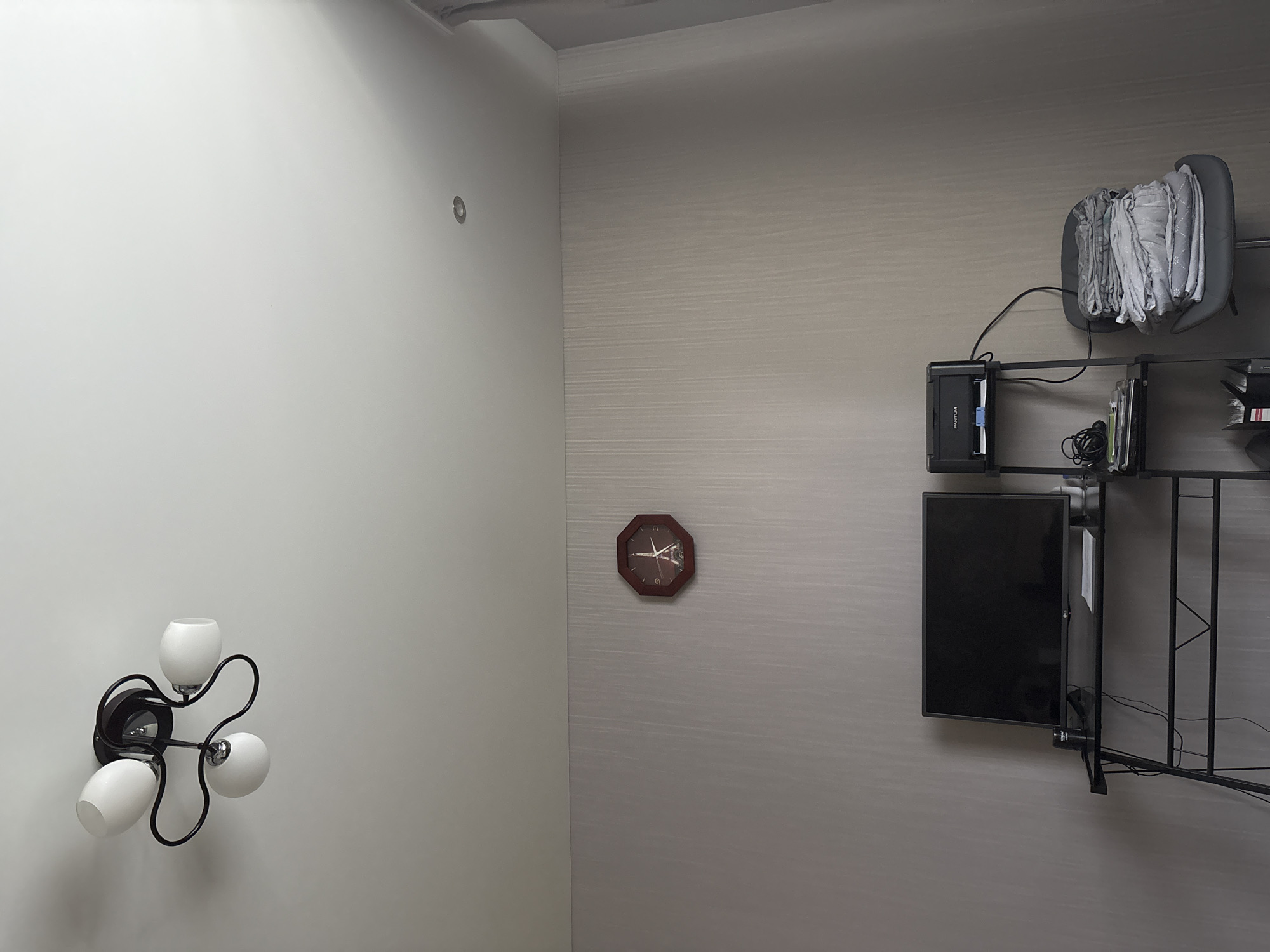The image depicts an indoor scene, likely a domestic setting, possibly in the city of Kinel'-Cherkasy, Russia. In the upper portion of the frame, a ceiling light fixture with three white glass shades on black metal arms is visible. Below this, a wall features a decorative octagonal clock with a dark wooden frame, displaying Roman numerals and a central image of the Eiffel Tower. Further down, a black metal desk or shelf unit holds a black flat-screen television on the left. To the right of the television, a small black printer is positioned on a higher shelf. To the right of the printer, a gray office chair is stacked high with folded gray linens, possibly towels or bedding. The walls appear to be covered in textured wallpaper. The overall lighting suggests an interior environment with ambient light. There are no people or animals present in the scene.