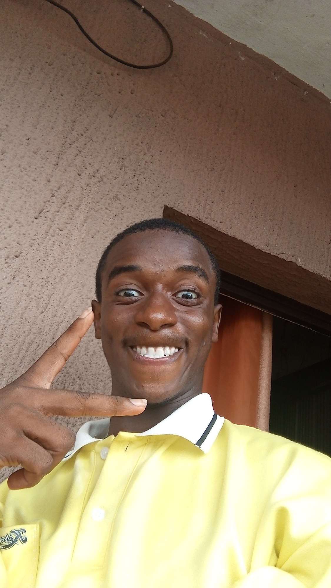 A young Black man is captured in a close-up, chest-up portrait, smiling broadly and looking directly at the camera. He wears a bright yellow short-sleeved polo shirt with a white collar accented by a thin black stripe. A small, illegible logo or design is visible on a pocket over his left chest. His expression is joyful and playful, characterized by a wide, open-mouthed smile revealing white teeth, and wide, bright eyes. His left hand is raised to the side of his head, with his index and middle fingers extended in a V-sign gesture, pointing towards his temple.
The setting appears to be an indoor or sheltered outdoor space during the daytime, indicated by bright, natural lighting. The background features a textured, light brown or beige wall, resembling stucco or rough plaster. In the upper left corner, a black electrical cable loops across the wall. To the right of the man's head, a dark brown, possibly wooden, doorframe or structural beam is visible. No discernible text is visible on his shirt or in the background beyond the unreadable logo. The overall atmosphere conveyed is one of cheerfulness and immediacy.
