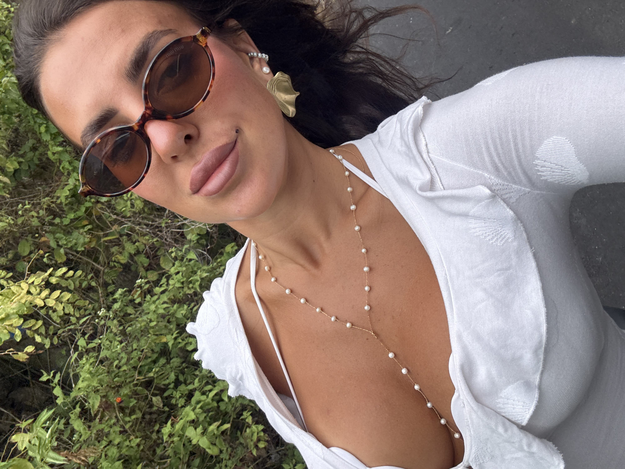 A woman, wearing a white top and a pearl necklace, is posing outdoors with lush greenery behind her. She is also wearing sunglasses and has bold makeup. The image captures a close-up view of her upper body and face, with a shallow depth of field that blurs the background.
