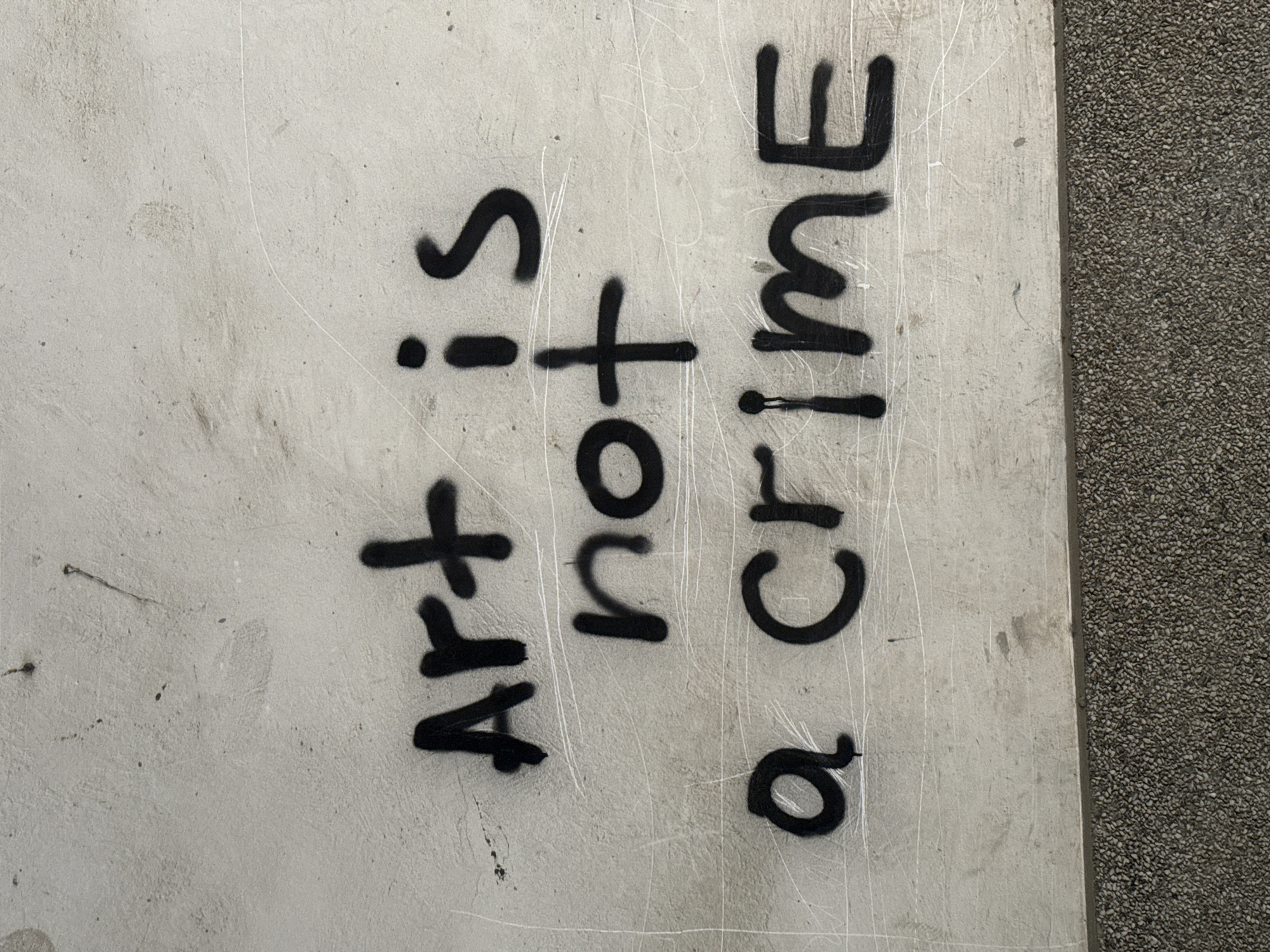 A spray-painted message reads "Art is not a crime" in black lettering on a light grey concrete wall. The wall is marked with scratches. A pebbled surface runs along the right edge of the frame, suggesting it is the edge of a sidewalk or road. The location is not evident from the image.