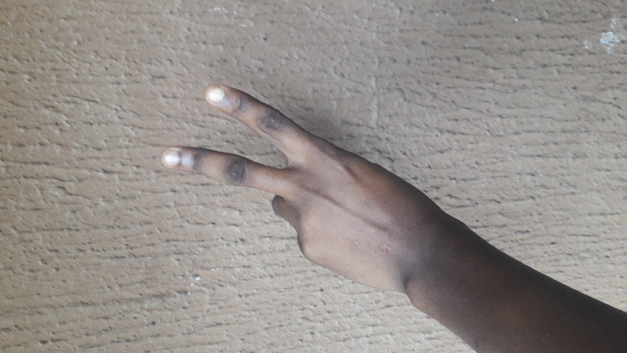 A close-up shot depicts a dark-skinned human hand positioned against a tan textured background. The hand is making a "peace" or "victory" sign with the index and middle fingers extended. The location in the city of Nkpor, Nigeria is not discernible from the visual data.