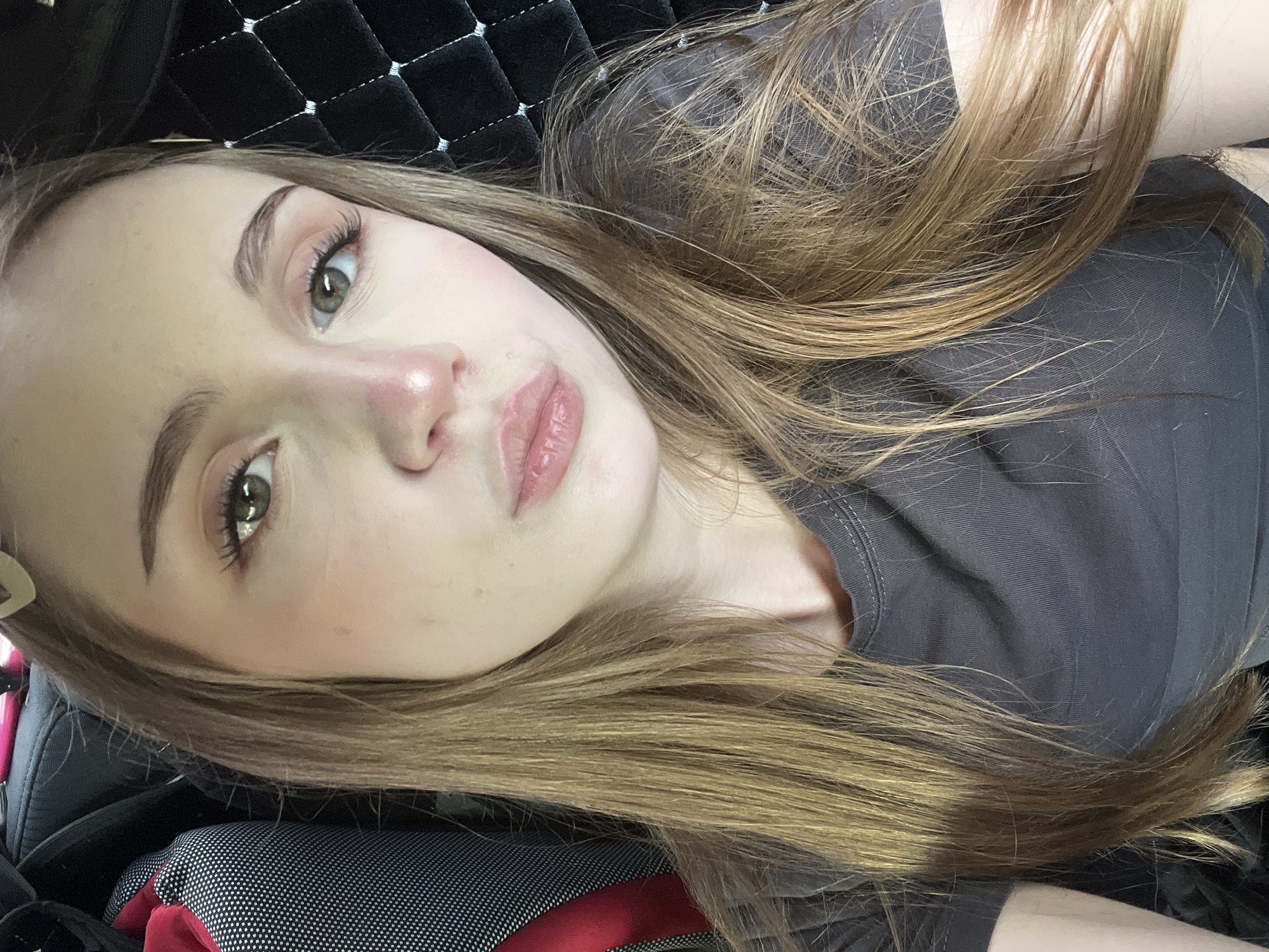 The image is a close-up selfie of a young Caucasian woman with long, straight, light brown hair. She is wearing a dark grey t-shirt. Her skin is fair, with visible blush on her cheeks and the tip of her nose. Her eyes are light green, and she has long eyelashes. Her lips are full and appear to be wearing a light pink lip gloss. She has a heart-shaped hair clip visible in her hair. The background appears to be the interior of a vehicle, with a textured black material on the right and a patterned fabric on the left, possibly a seat or luggage. The lighting suggests an indoor or car interior setting. There is no indication of the specific location within Загородный, Russia.