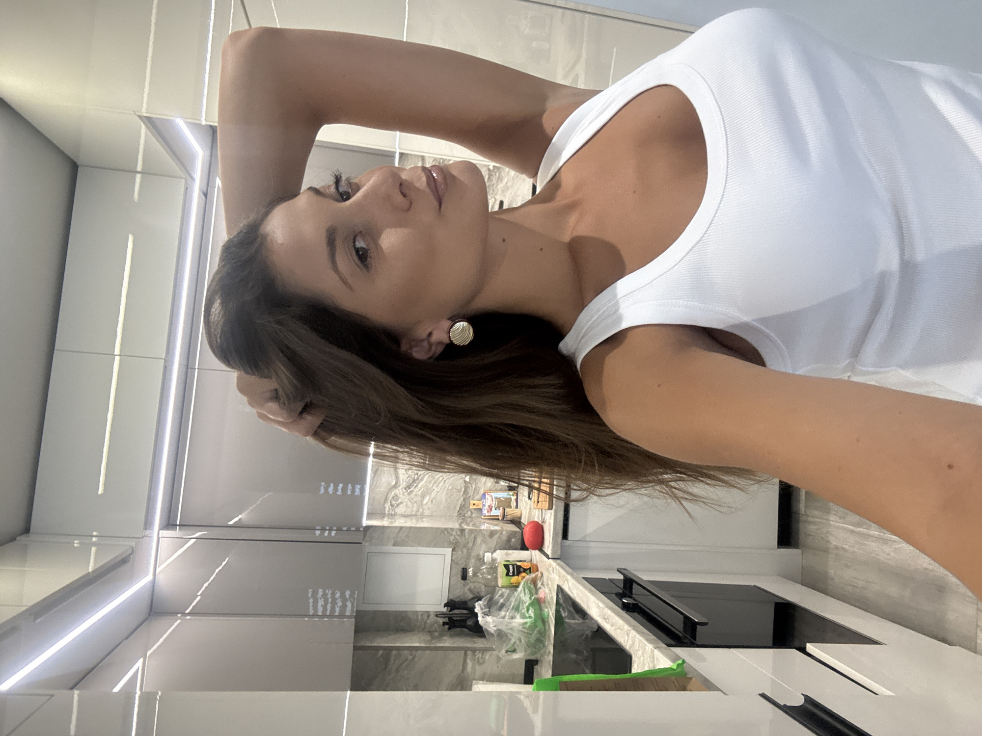A woman is posing for a selfie in a modern kitchen. The kitchen features white cabinets with integrated LED lighting, a grey backsplash, and a black stovetop. On the countertop, there is a plastic bag, a bottle of juice, and a wooden cutting board with a red item. The woman is wearing a white tank top and gold earrings. Her hair is brown and styled. No information suggests that the location is in the city of Burgas, Bulgaria.