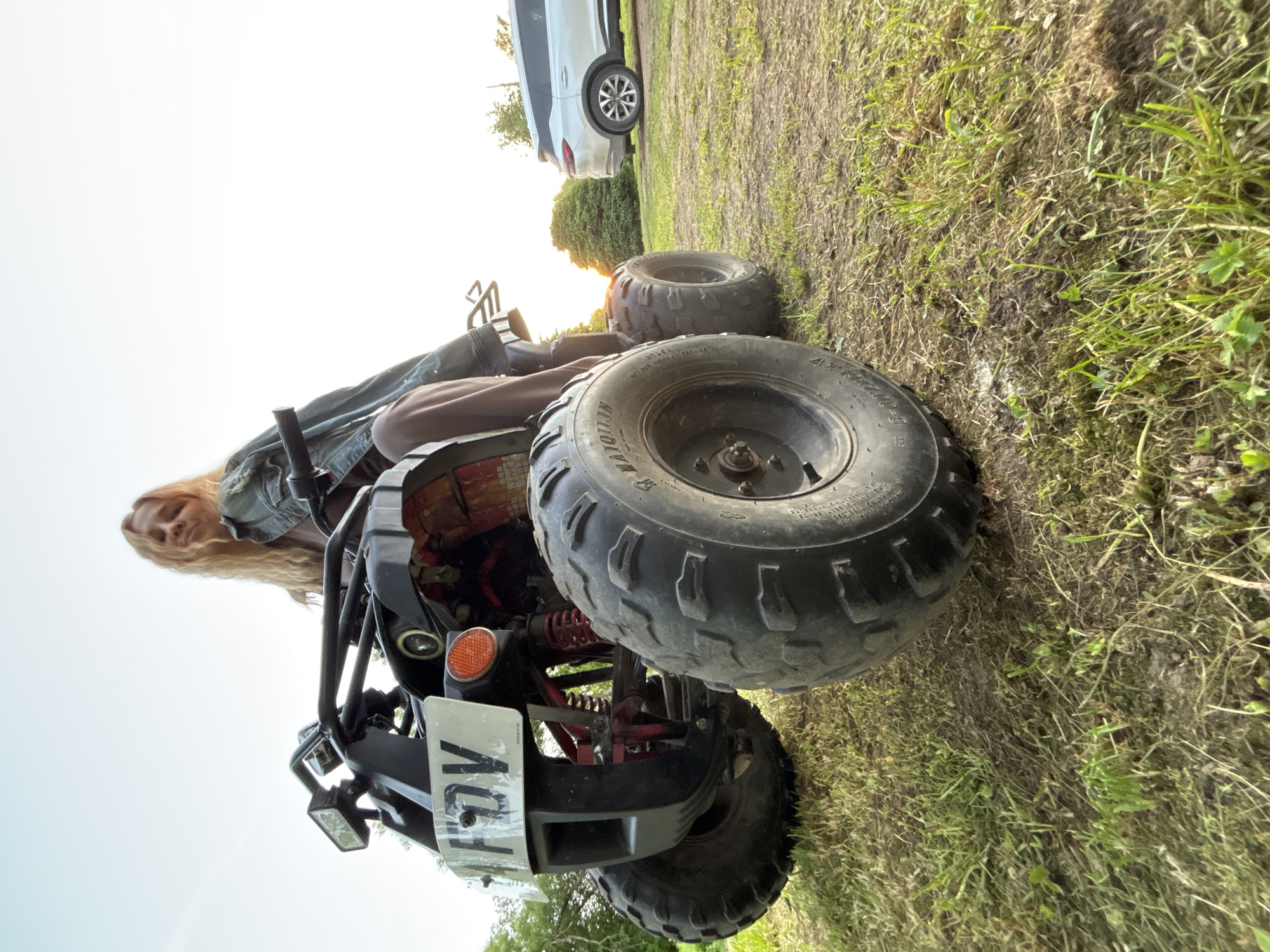 The image, captured in Rozhdestveno, Russia, presents a low-angle view of a blonde female seated on a black all-terrain vehicle (ATV) on an unpaved surface comprised of sparse grass and exposed dirt. The female subject, positioned towards the center-left, looks directly at the camera with a neutral expression, wearing a denim jacket and light-colored trousers, with her legs visible on the ATV. The front of the ATV is angled towards the lower-left, featuring a white license plate displaying the characters "F DV," a rectangular front headlight on the left, and a round orange reflector on the right. Red suspension springs are visible beneath the black frame. The large, knobby front tires, including the right front tire with "MAIQILIN AT19X7.00-8" embossed, are prominent in the immediate foreground. In the mid-ground, a dense green bush partially obscures a low-angle sun, casting a warm, bright light from the right. A white SUV is parked further in the background on the right side of the frame. The sky above is clear, transitioning from light blue to a bright, sunlit white on the right.
